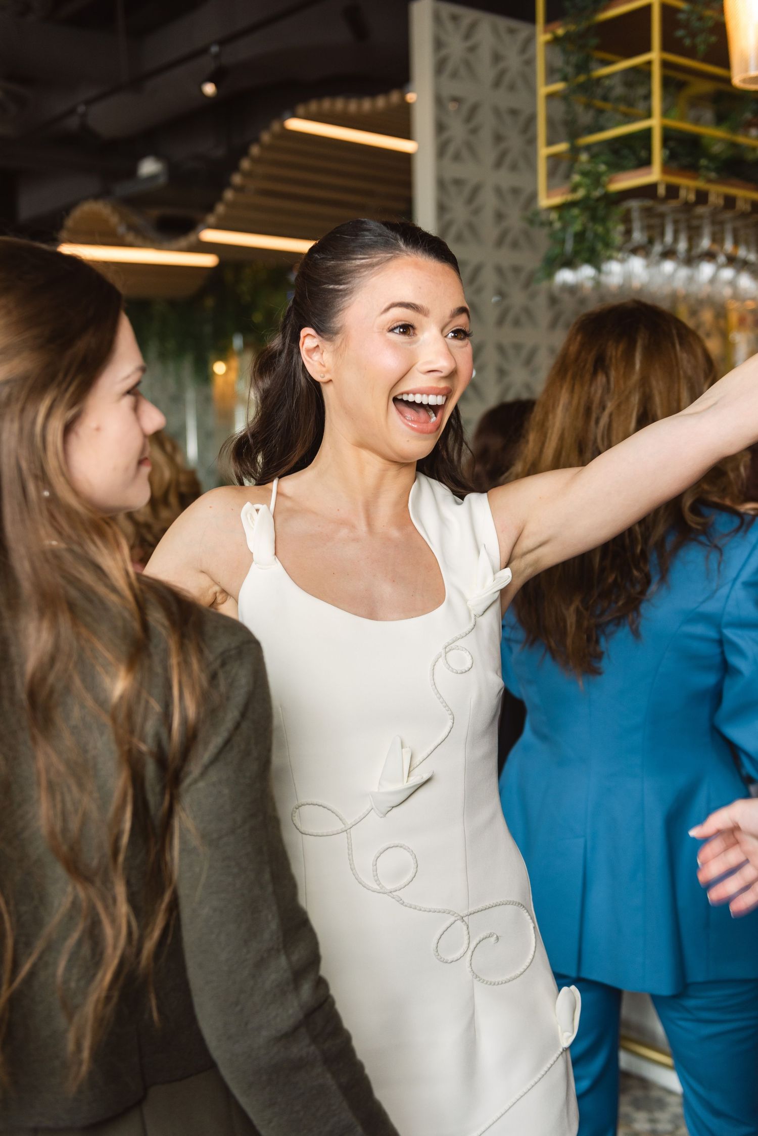 A joyful woman in a white dress laughs and dances with friends at an elegant indoor celebration.