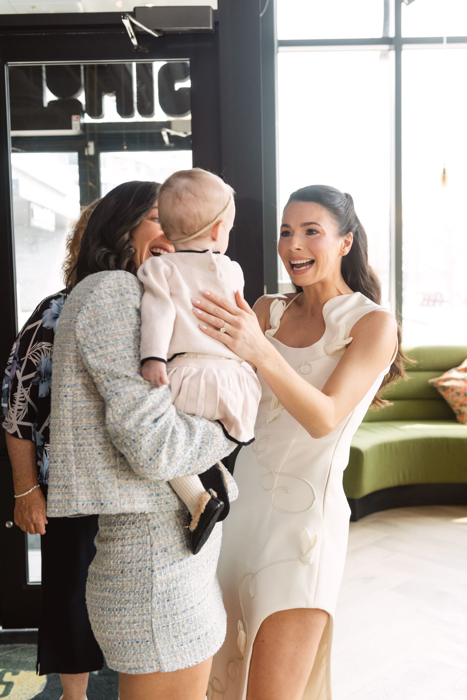 Two elegantly dressed women admire a baby in a pink outfit inside a stylish venue.