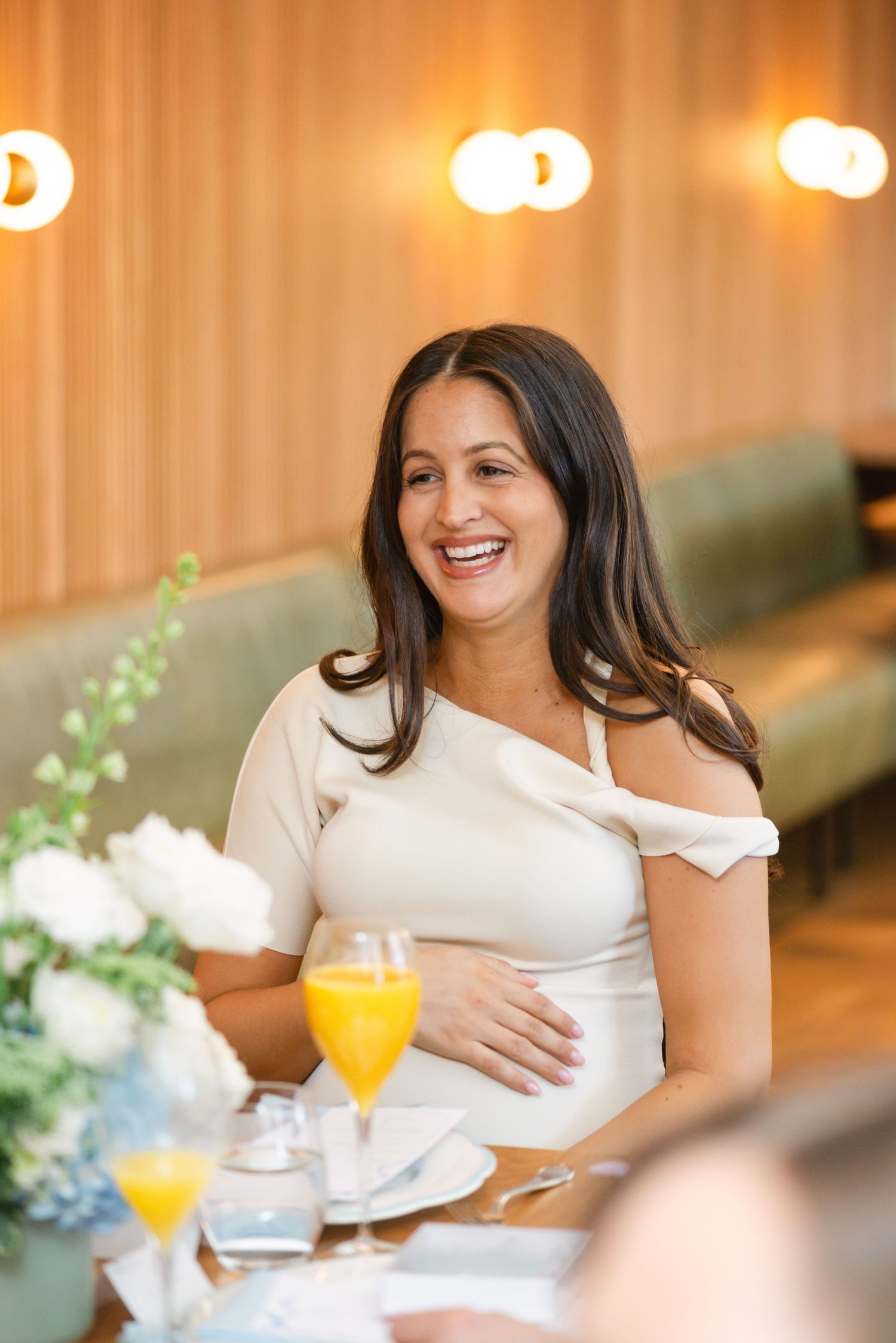 A woman in white dress sits at a table smiling and laughing during a celebratory event.