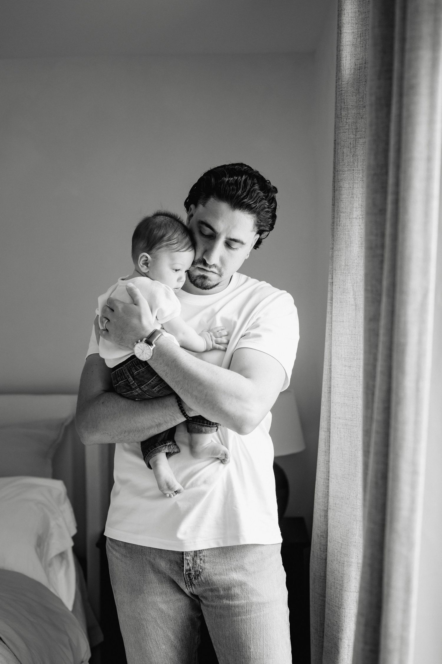 A tender black and white portrait of a parent cradling a sleeping baby while standing near a window.