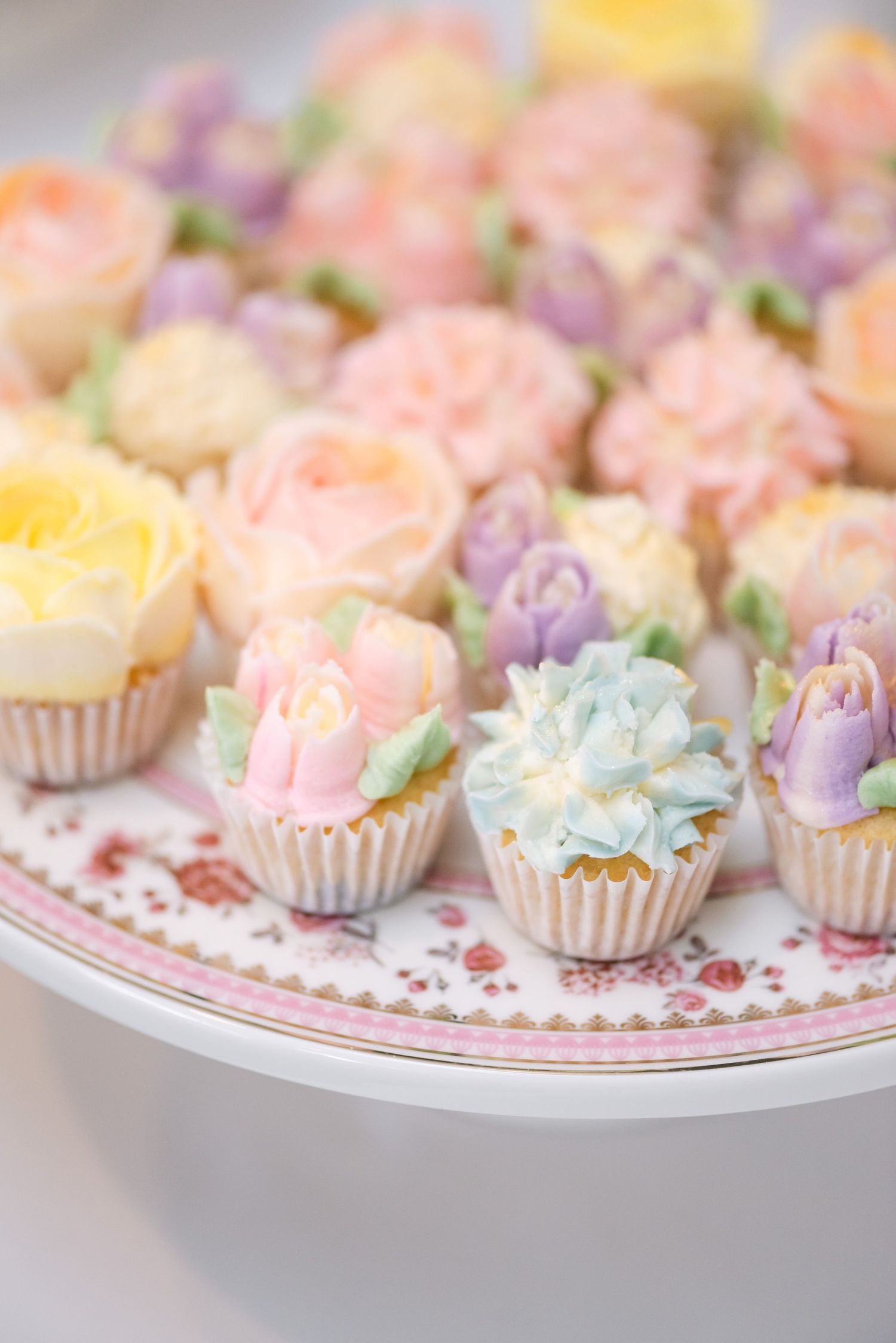 Assorted pastel floral cupcakes with buttercream flower designs on a decorative cake stand.
