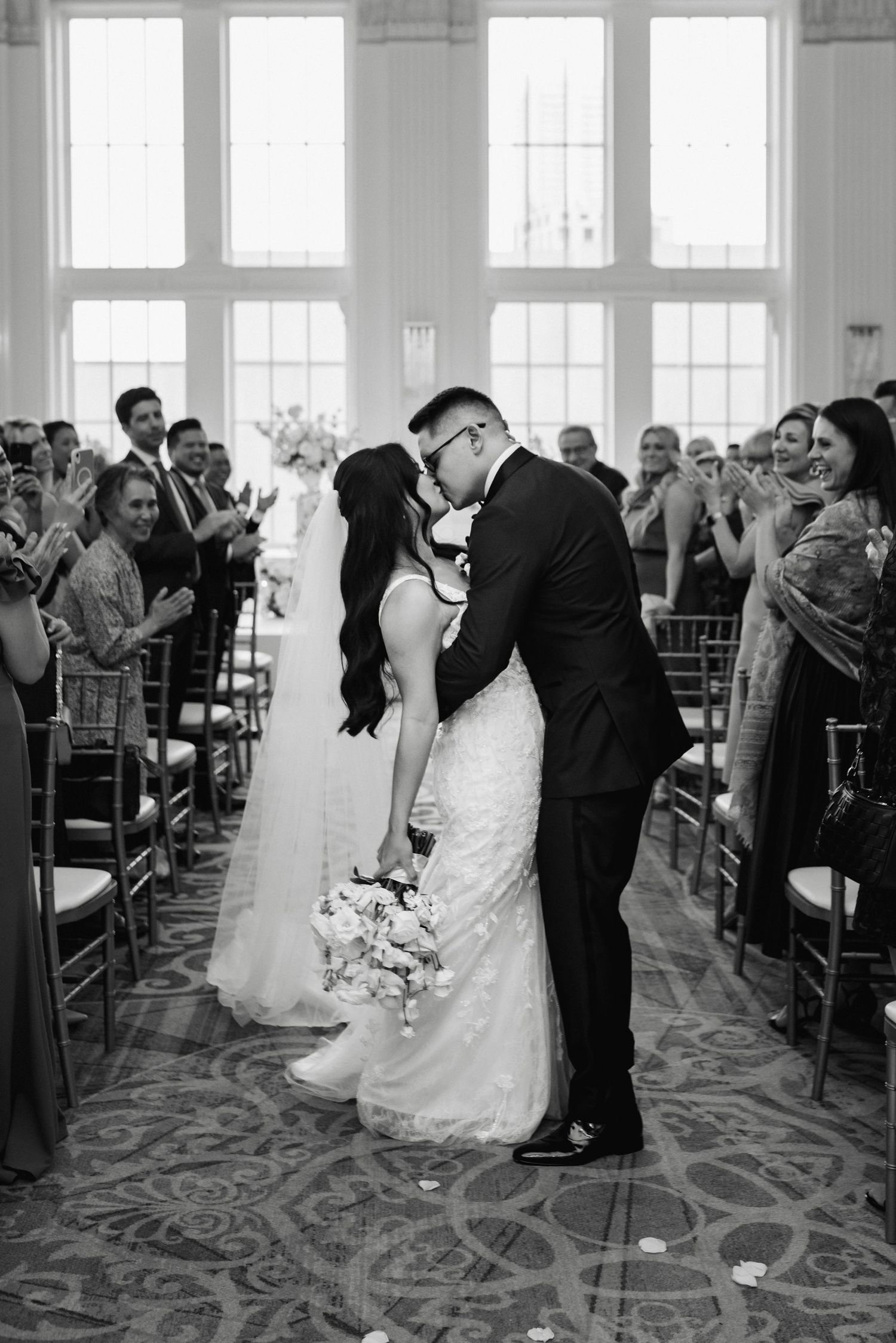 Wedding ceremony processional in an elegant ballroom with guests seated in wooden chairs.