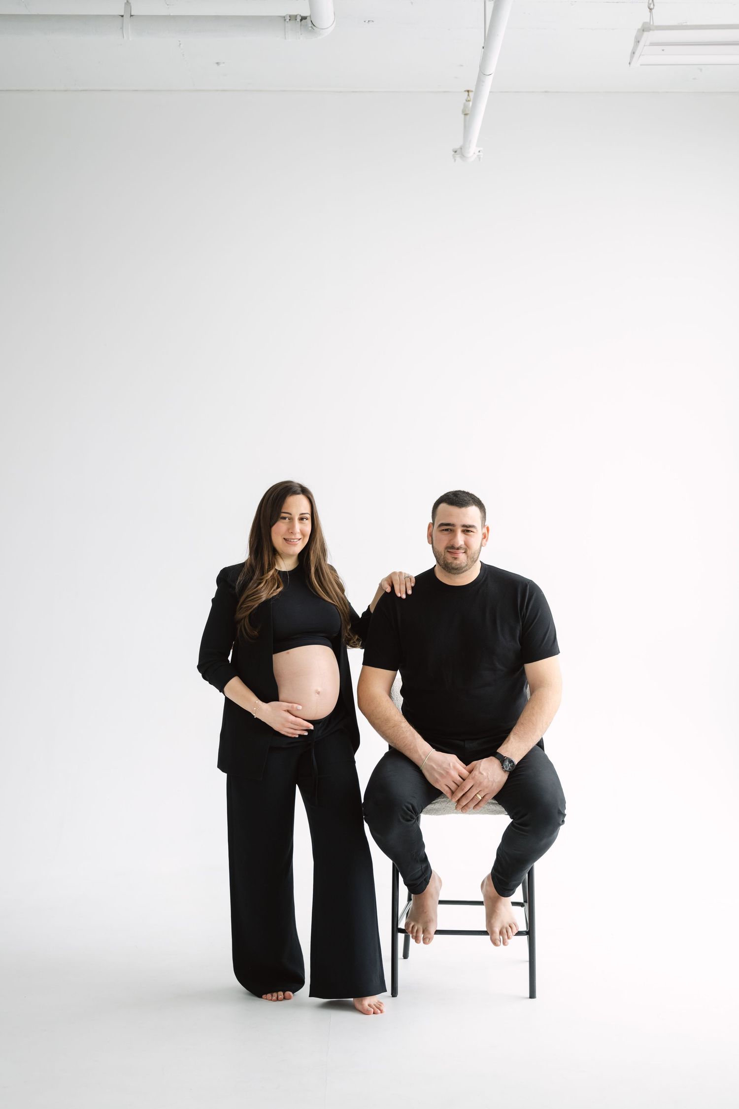 A couple in matching black outfits poses in a minimalist white studio setting during a maternity photoshoot.