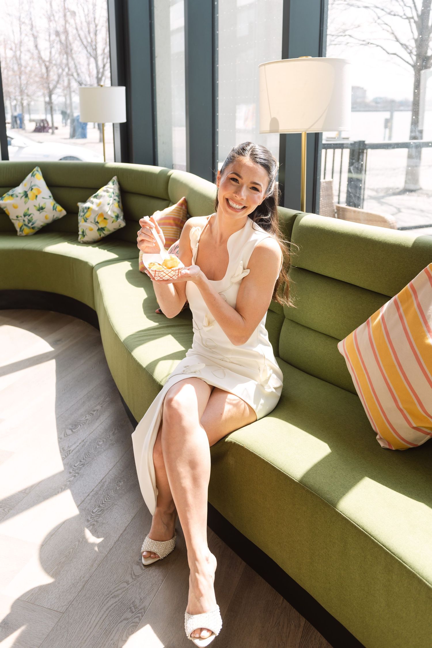 Happy bride in white dress sitting on green curved sofa holding a colorful citrus drink indoors.