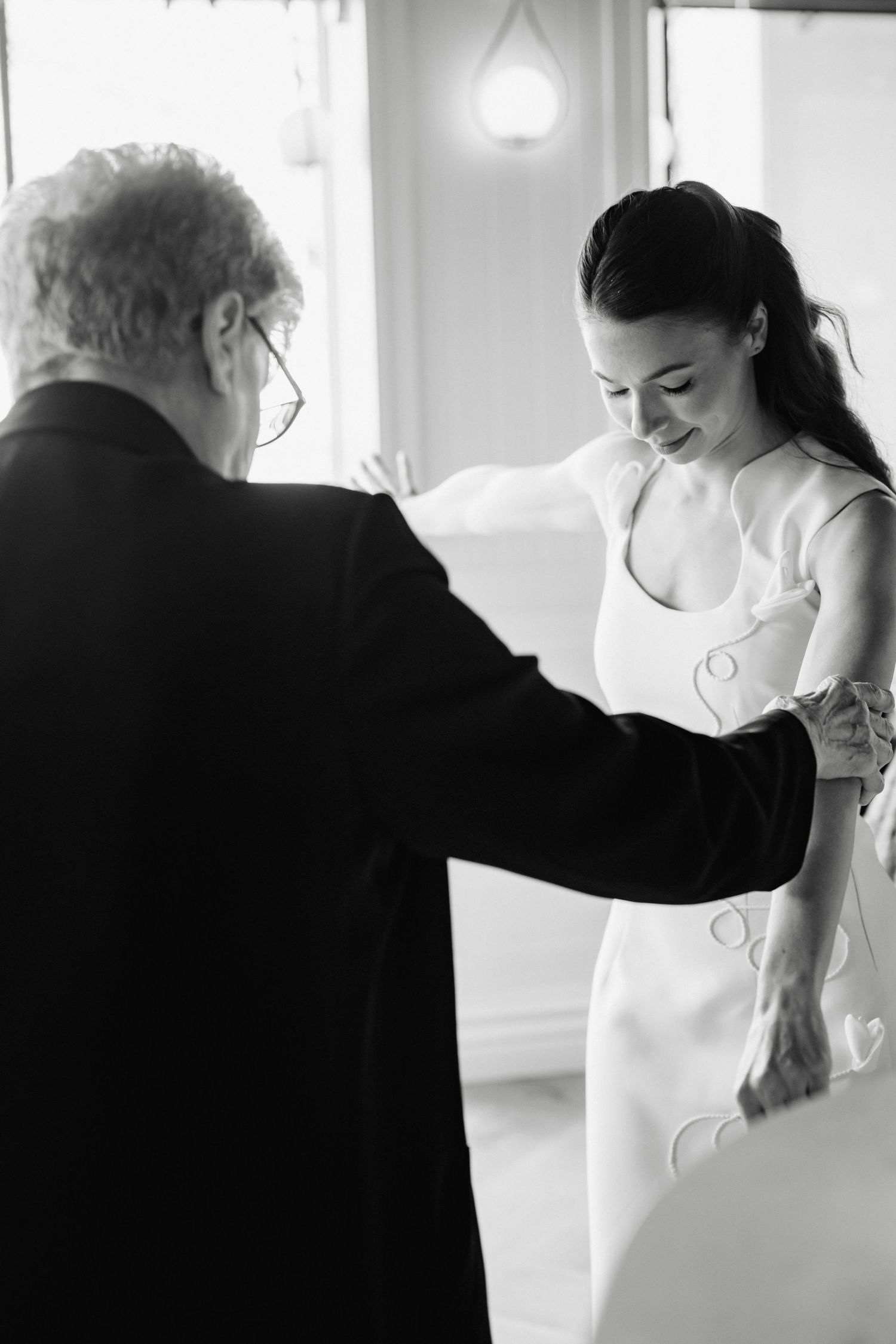 Black and white photo of a bride in white dress smiling as an elderly man helps her prepare for her wedding.