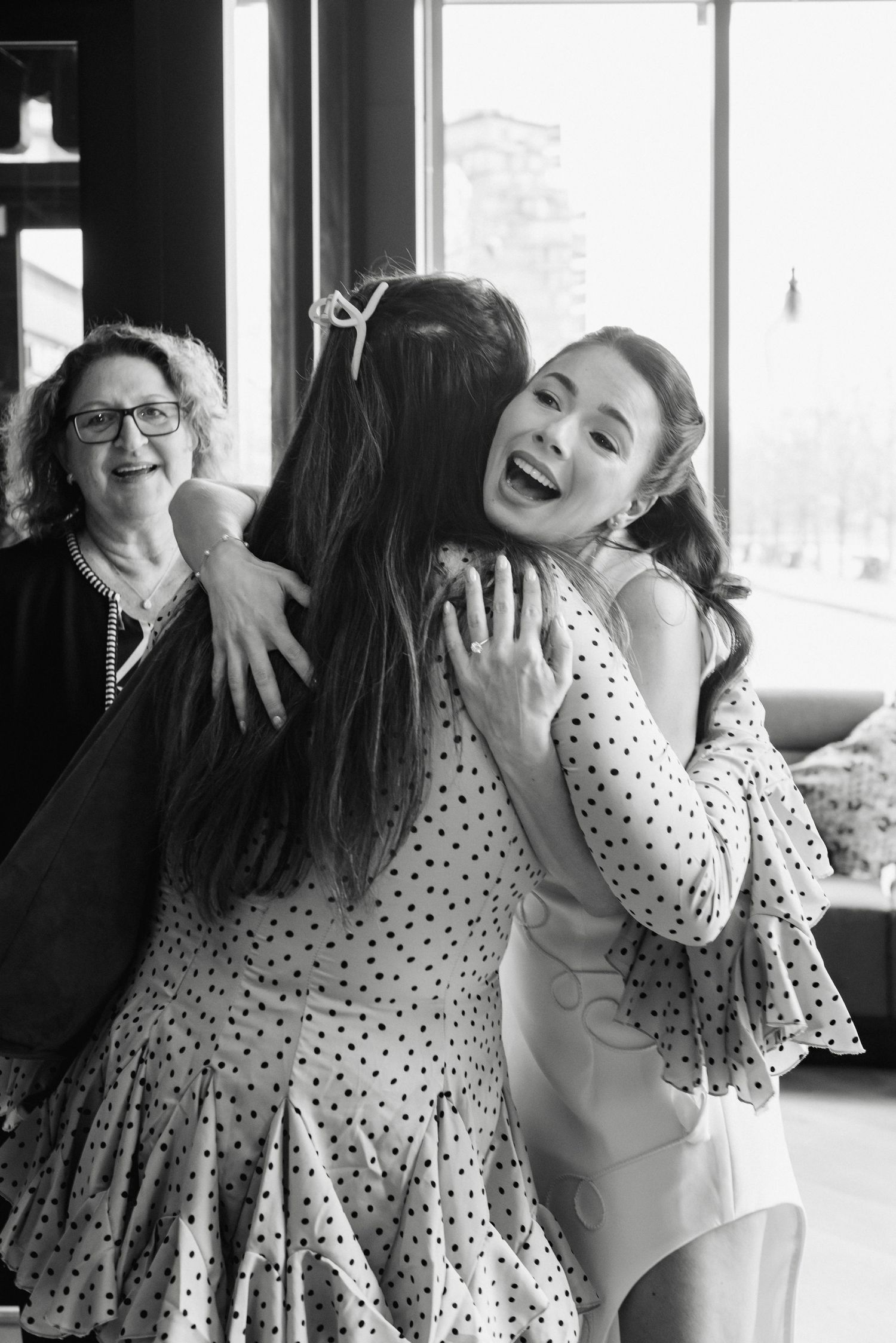 Two young women share a joyful hug while an older woman smiles warmly in the background, black and white photo.