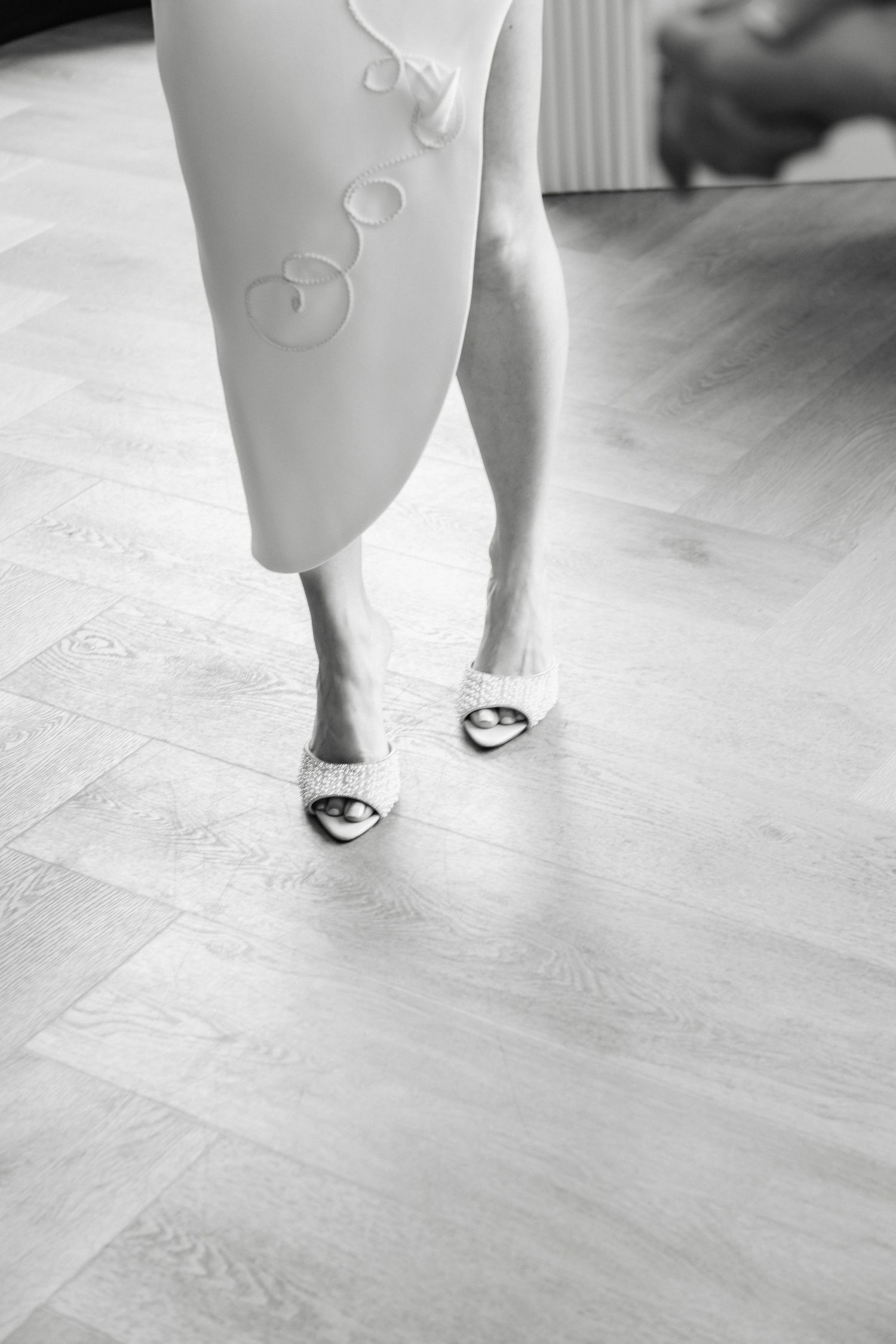 Close-up of a woman's feet in open-toe heeled sandals wearing a white dress on a light wood floor.