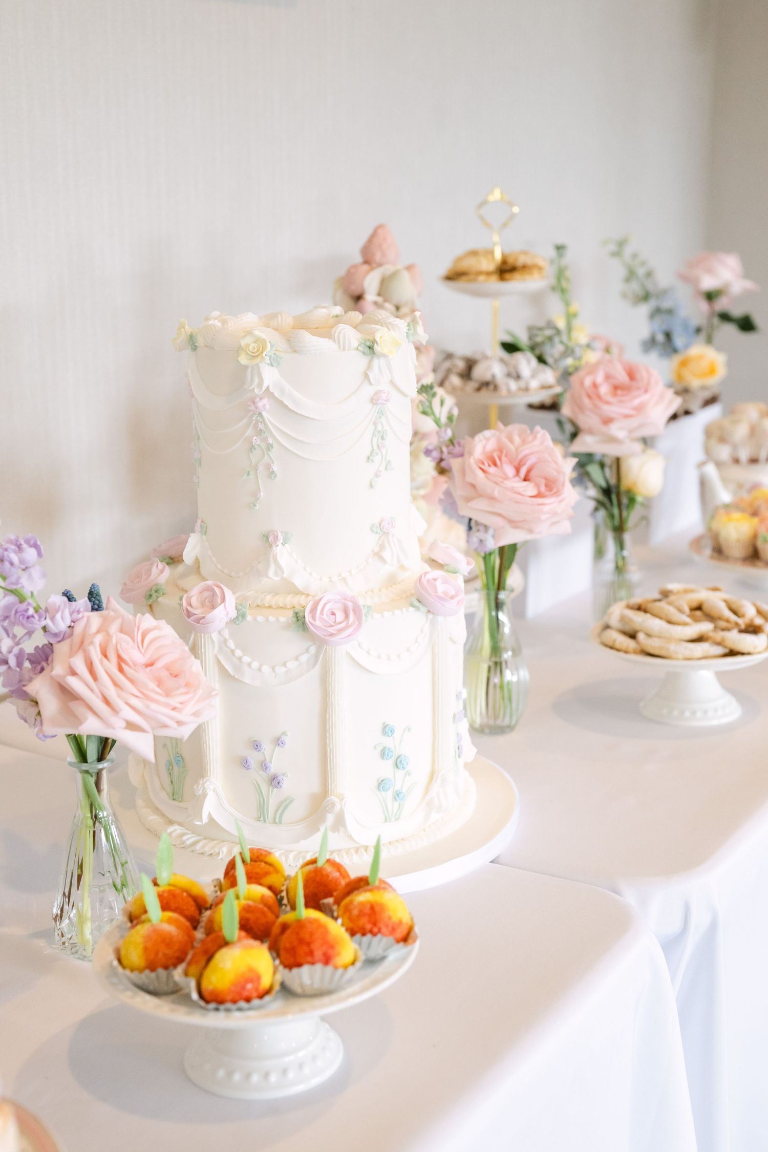 Elegant white two-tier wedding cake with pastel roses and floral decorations on a dessert table.