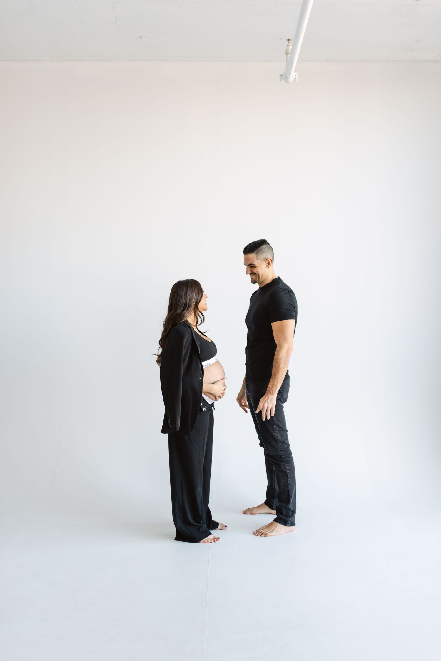 Full-length maternity photos of couple in black attire against white backdrop.