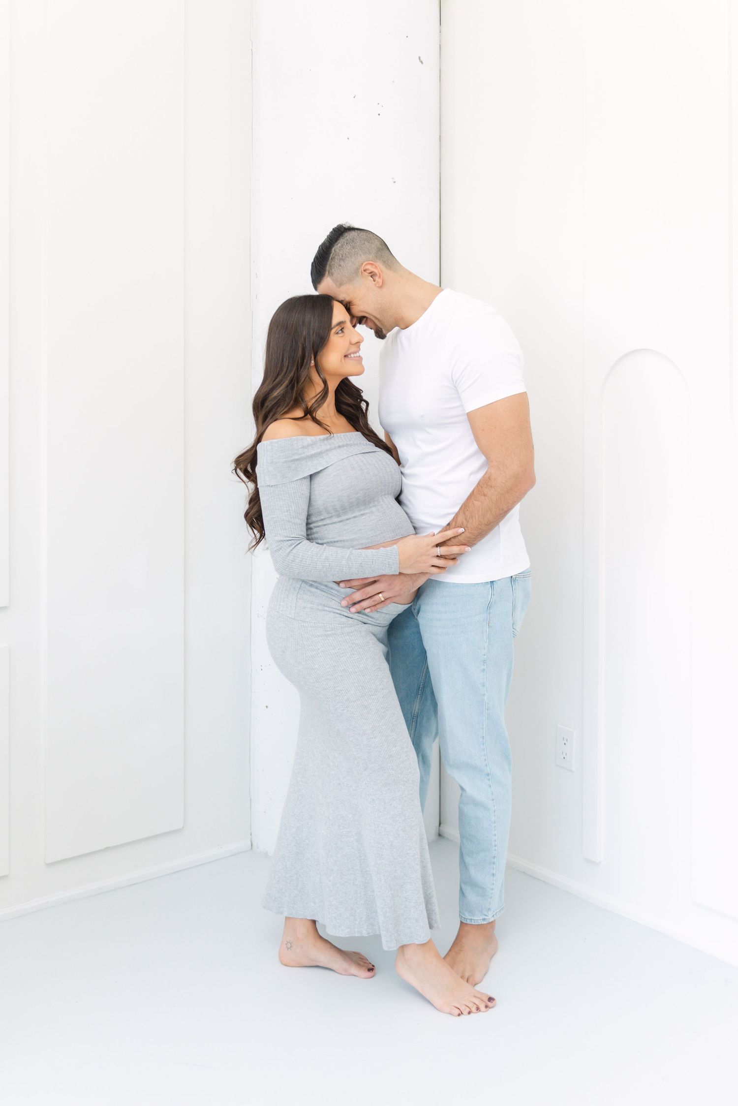 Maternity session with couple in casual attire against white wall.