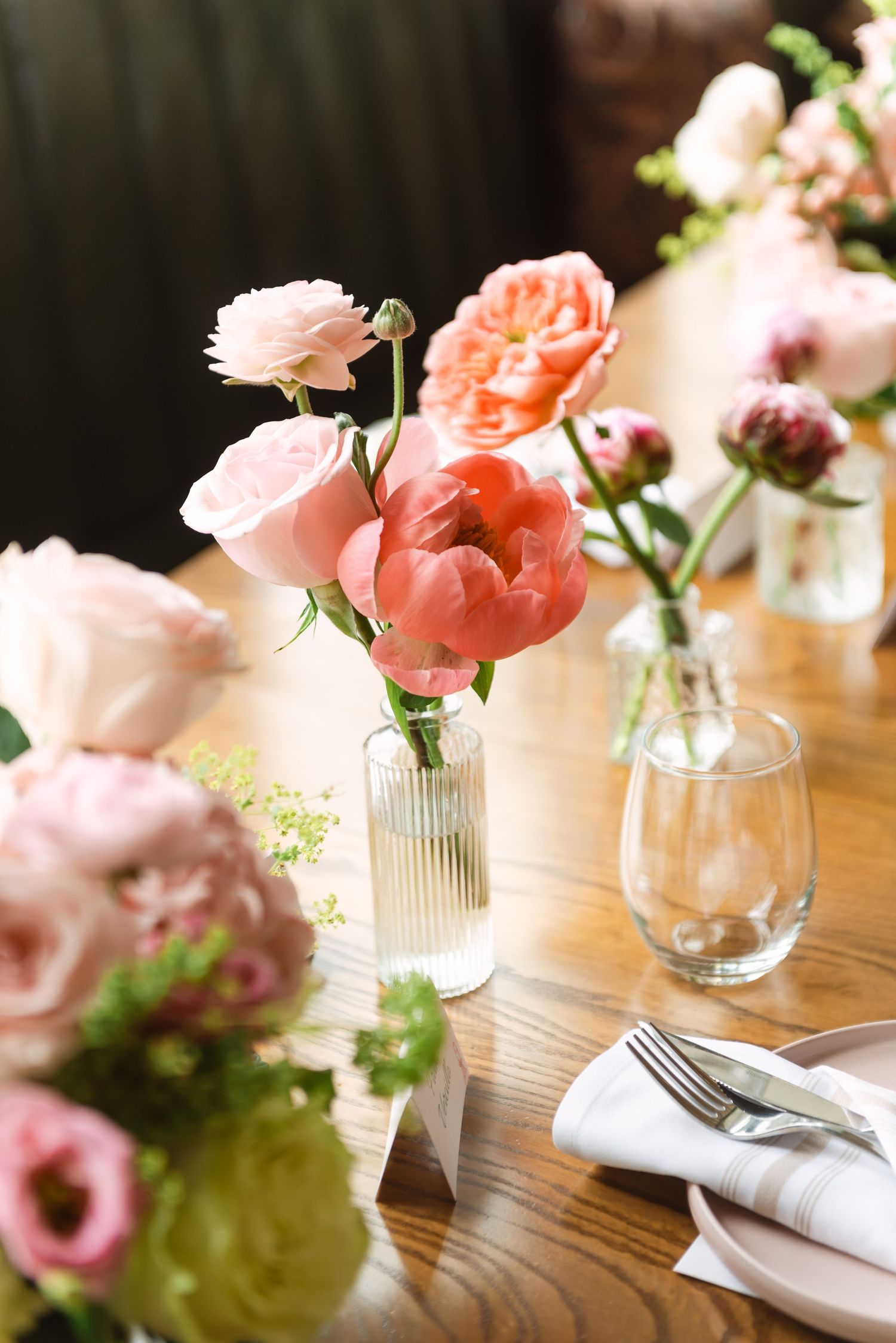 Elegant table setting with pink roses and coral tulips in glass bud vases on a wooden table.