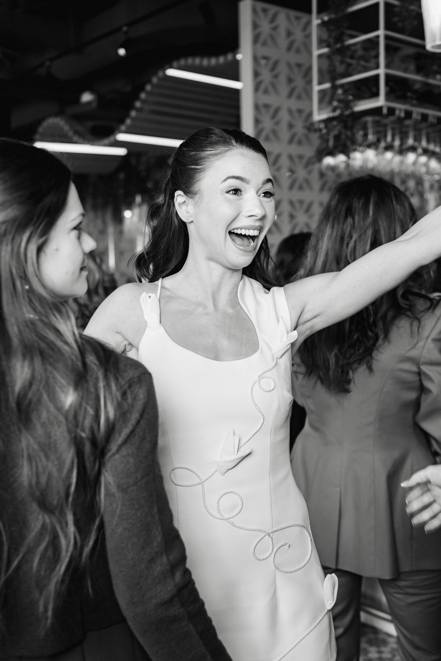 Black and white photo of a joyful woman in a white dress dancing and laughing at a party with friends.