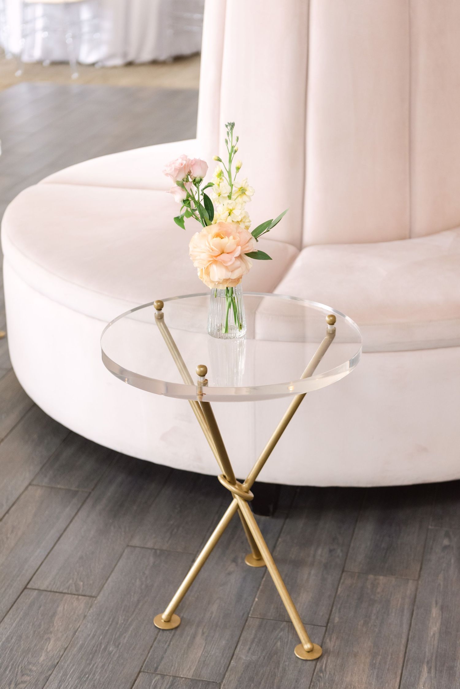 Gold tripod side table with glass top holding pink flowers beside a curved blush velvet sofa at an elegant event.