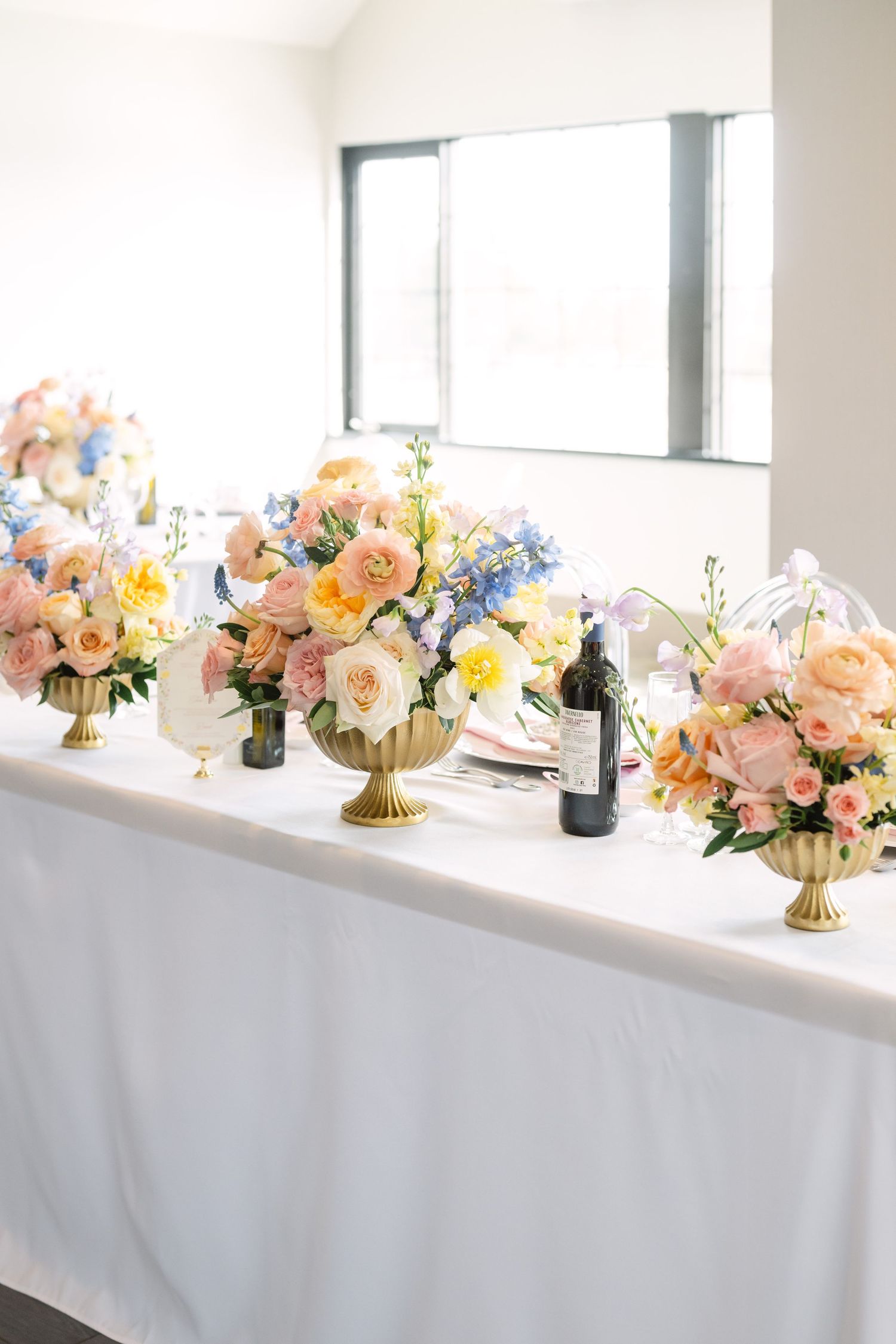 Elegant wedding head table with pastel floral arrangements in gold vases, featuring pink, blue, and yellow blooms on white linen.
