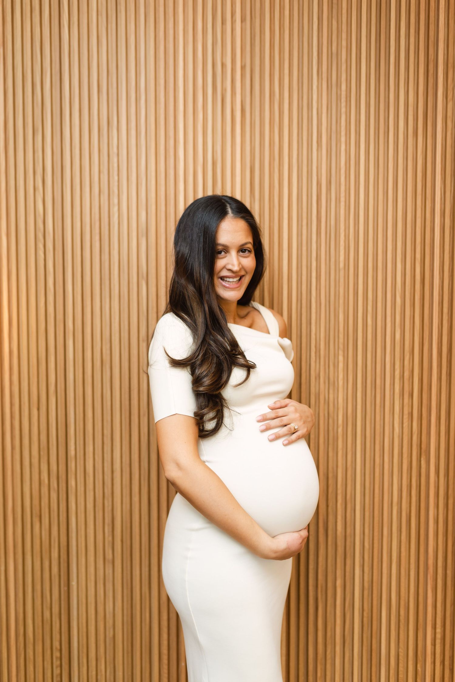 A pregnancy progression photo series showing a growing baby bump against a wood paneled wall in white dress.