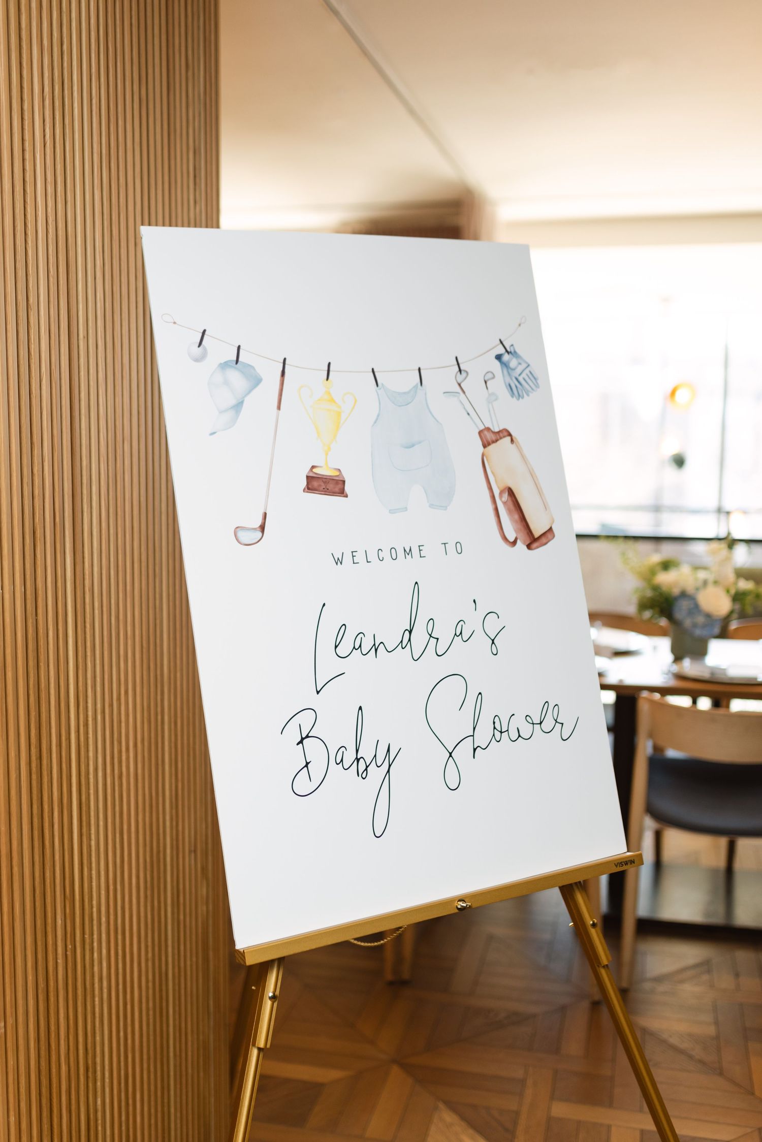 A white welcome sign for a baby shower stands on a gold easel, decorated with watercolor baby clothes on a clothesline.