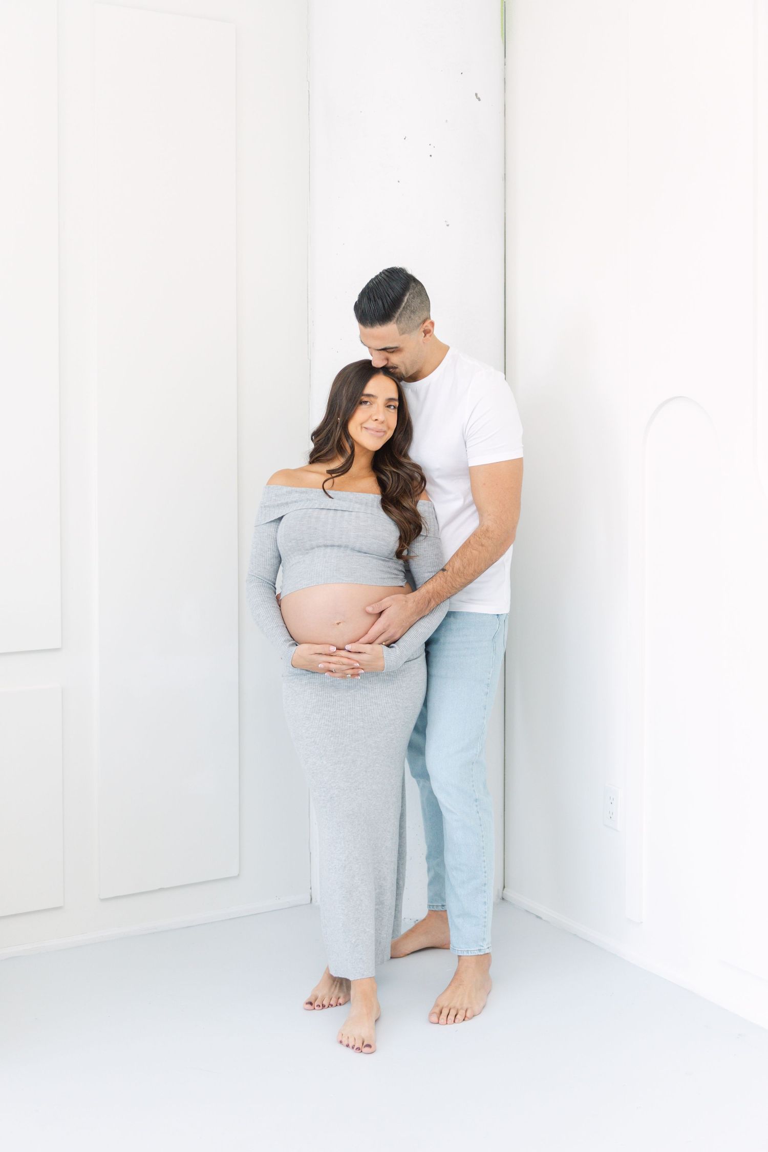 Modern maternity photography with couple posing against white walls in minimalist setting.