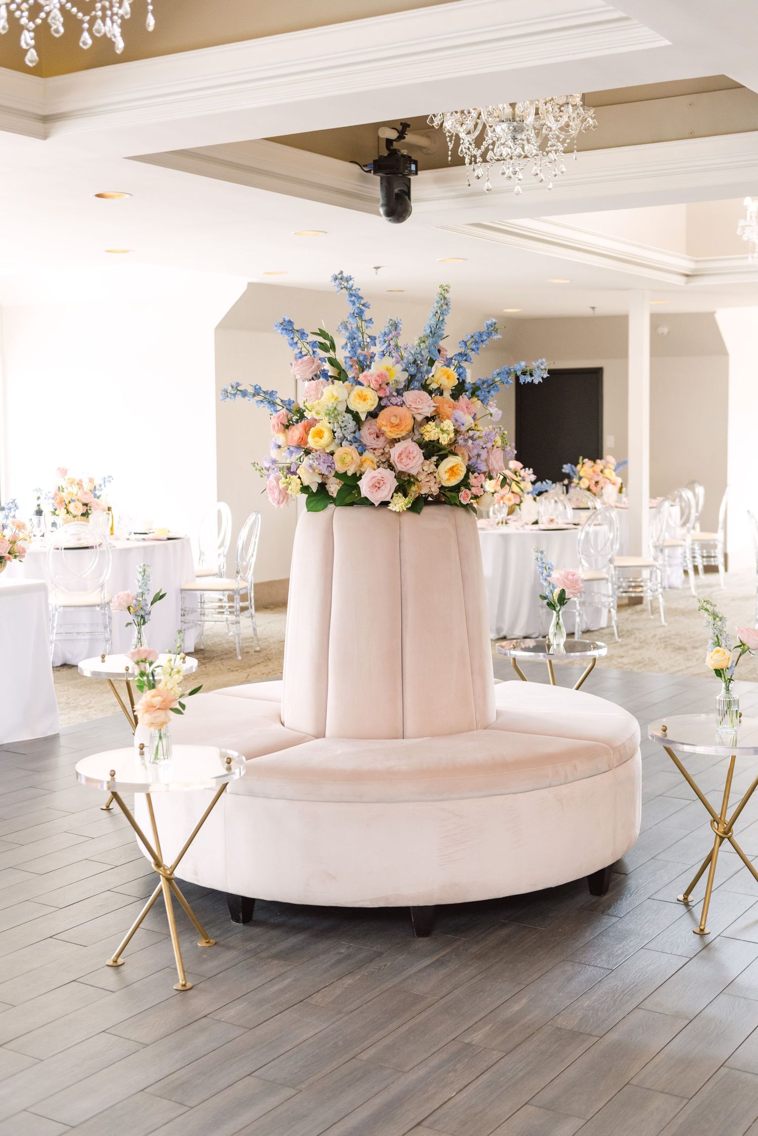 Elegant wedding reception with a blush circular velvet sofa, colorful floral arrangement, gold accent tables, and crystal chandeliers.