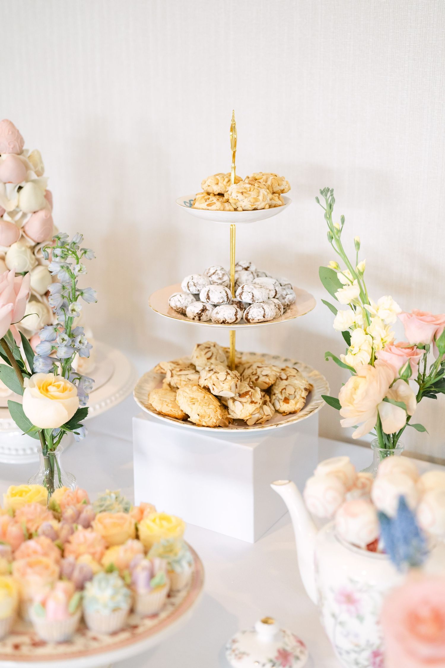 Elegant three-tier dessert stand with cookies and pastries surrounded by pastel floral arrangements at bridal shower.