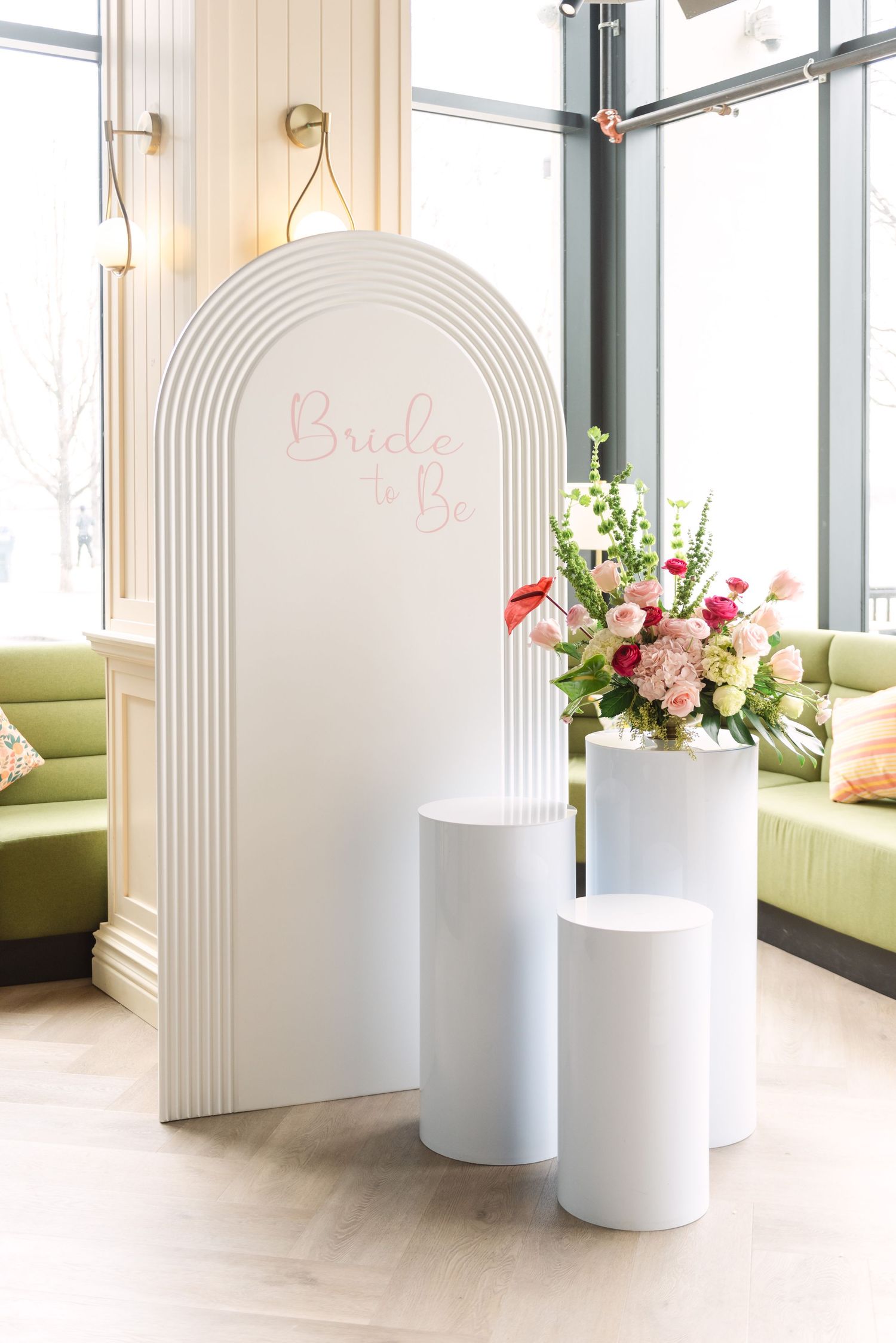 Elegant bride-to-be bridal shower setup with white arch backdrop, pedestal stands, and pink floral arrangement in a bright venue.