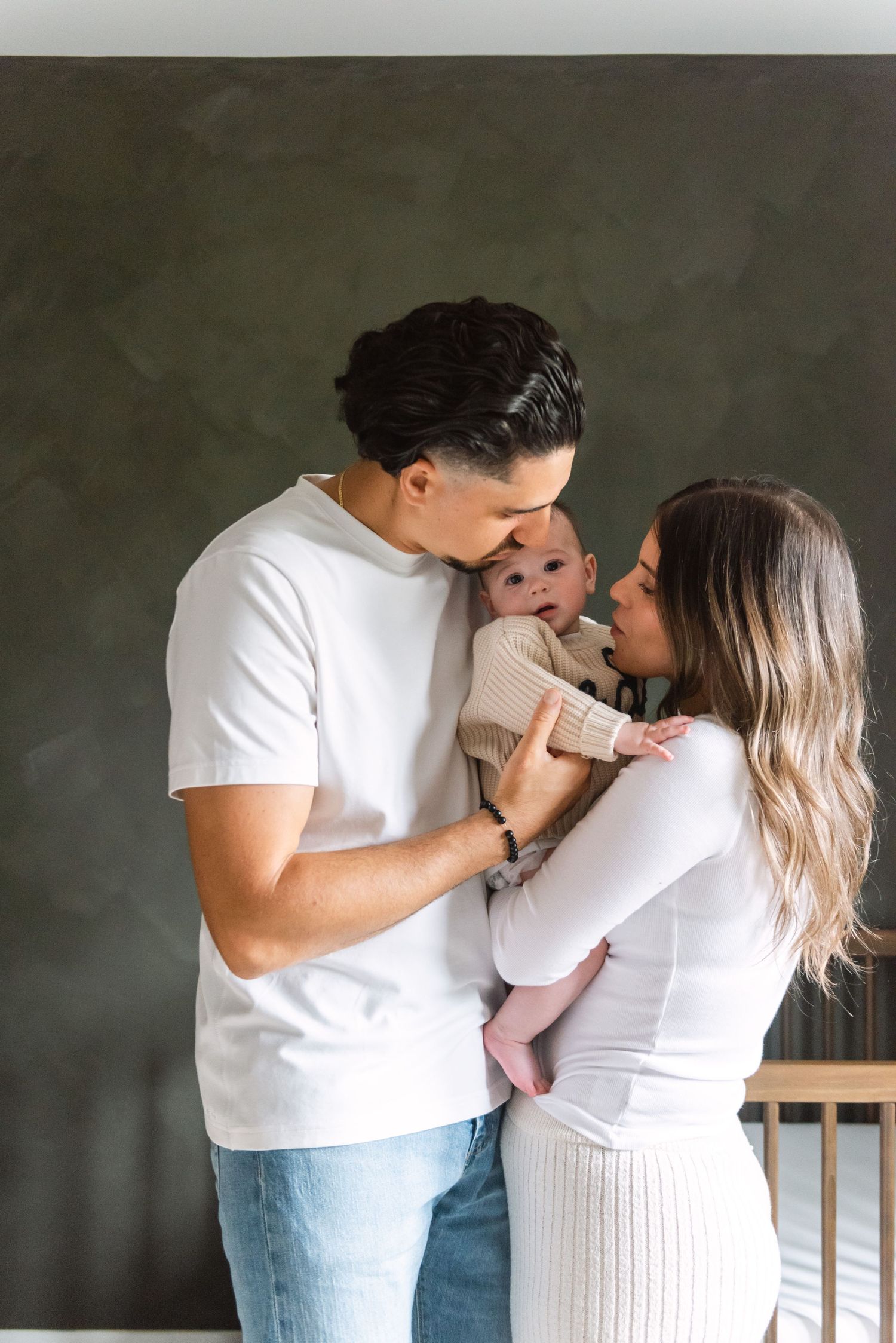 A couple in white outfits sharing an intimate moment with their baby against a dark gray background.