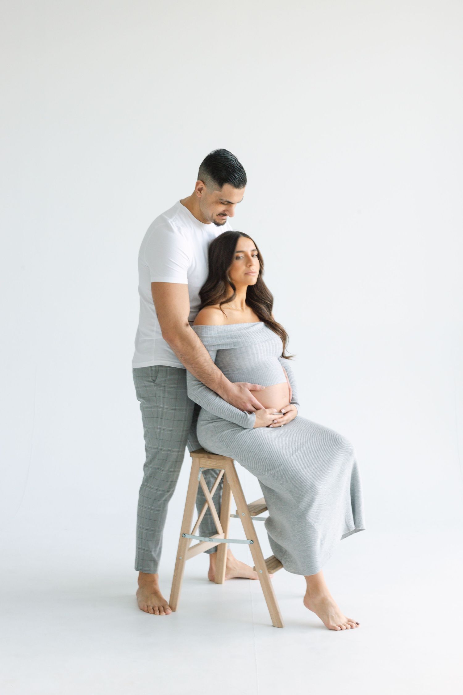 Couple shares tender moment during maternity photoshoot with wooden stool in minimalist studio.