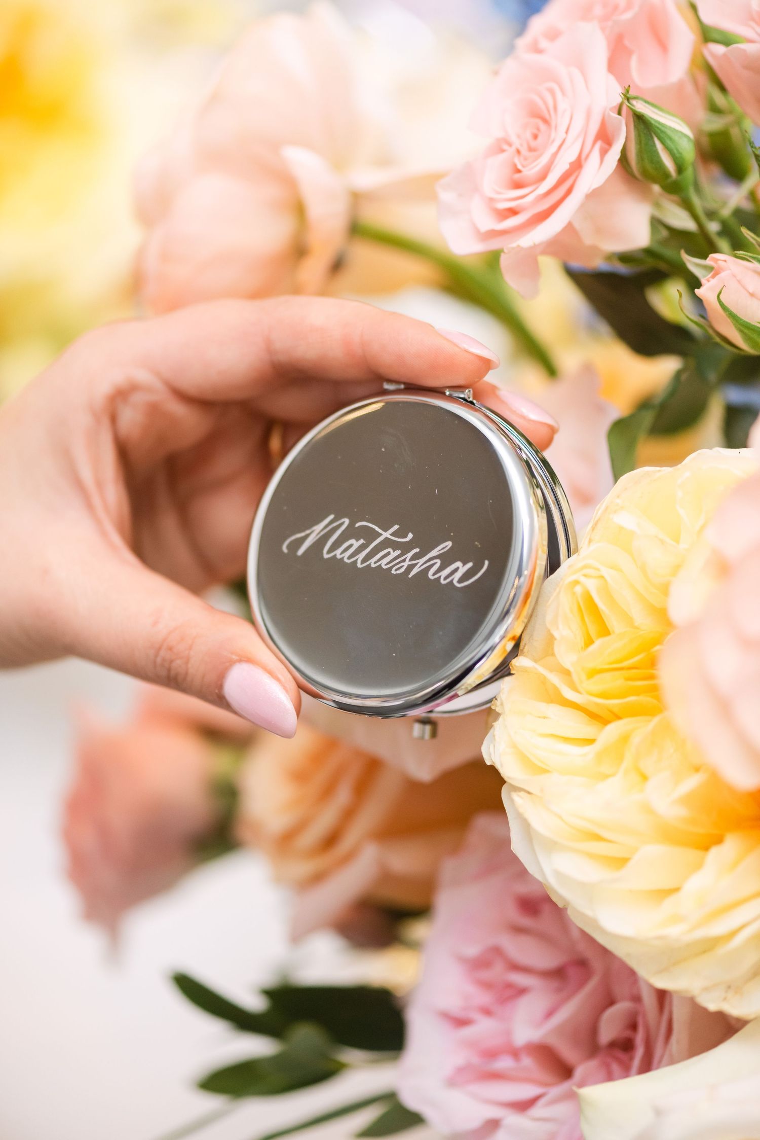 Hand holding engraved silver compact mirror with name Natasha surrounded by pink and yellow roses.