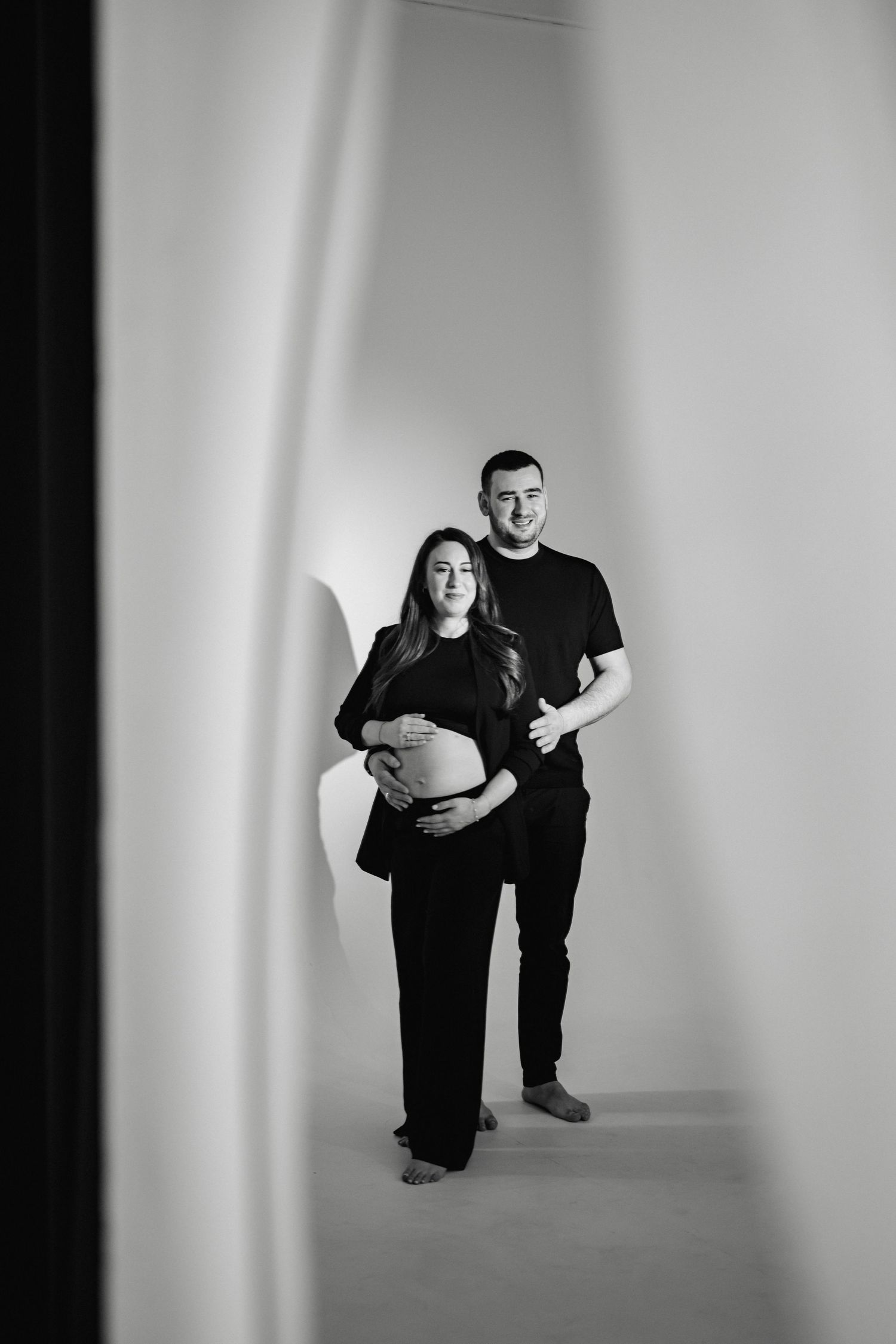Black and white maternity photo showing an expecting couple in dark clothing posing against a light studio backdrop.