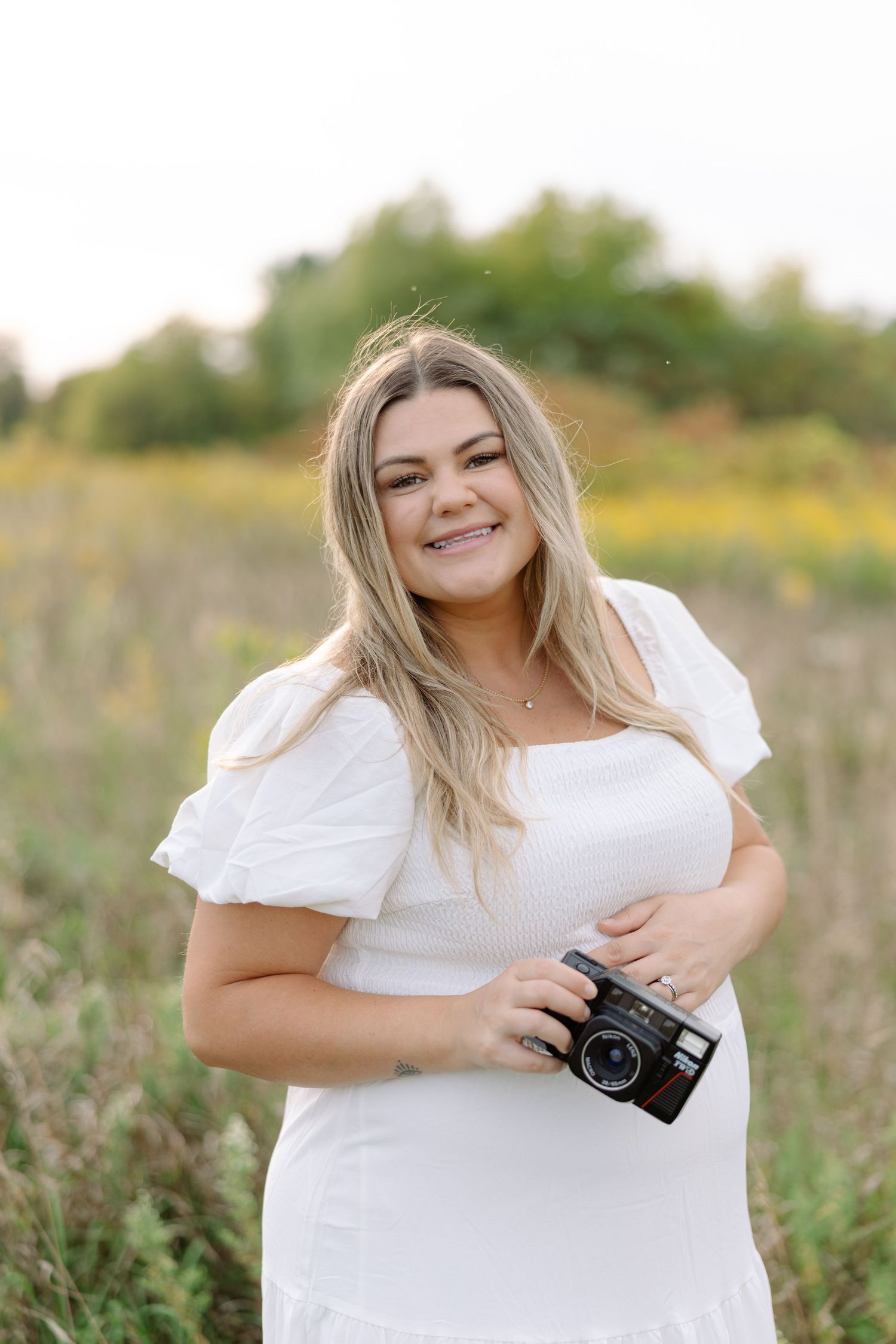 Woman in white dress poses with vintage camera in flowering meadow.