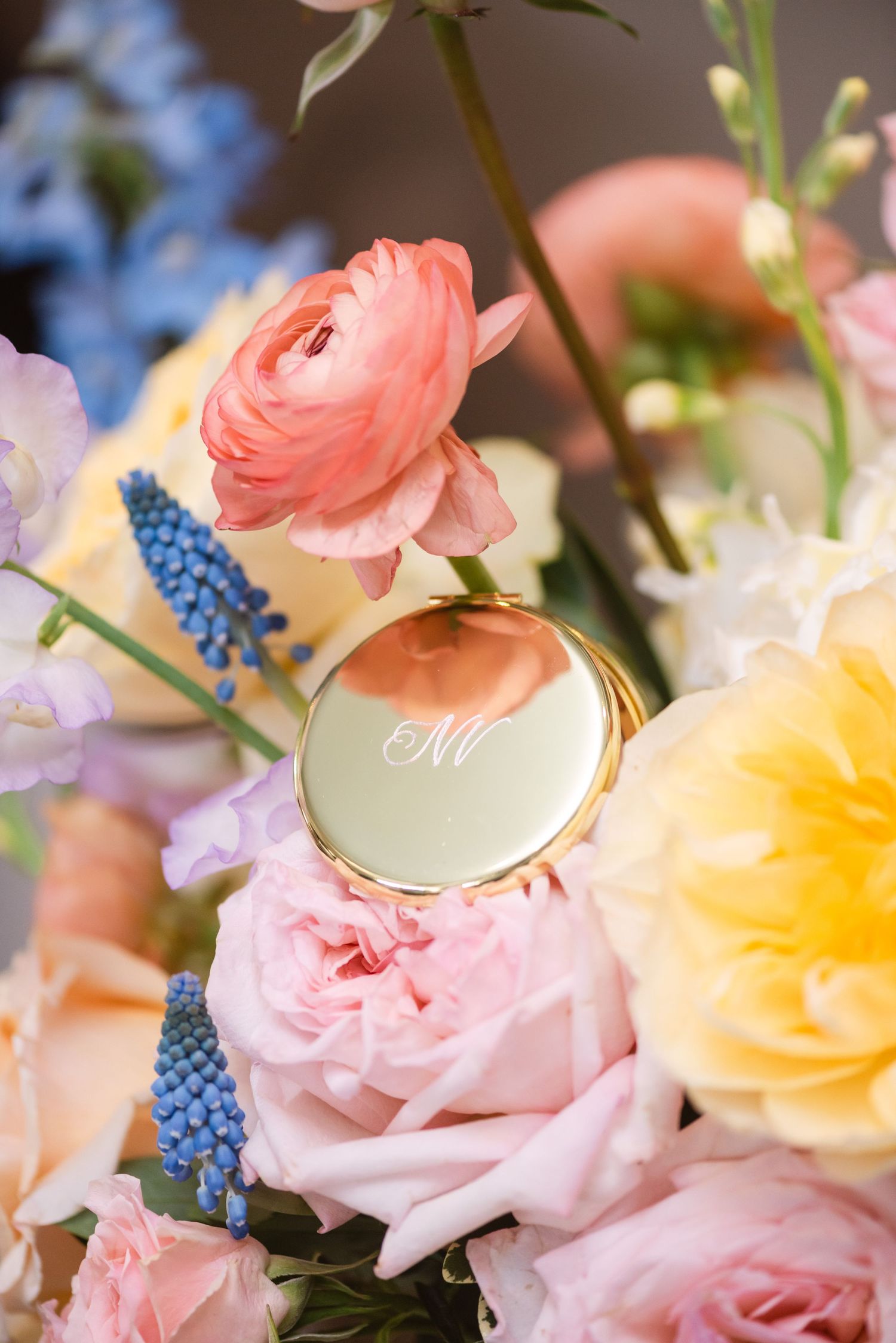Gold monogram compact mirror nestled among pink ranunculus, blue muscari, and pastel roses in a spring floral arrangement.