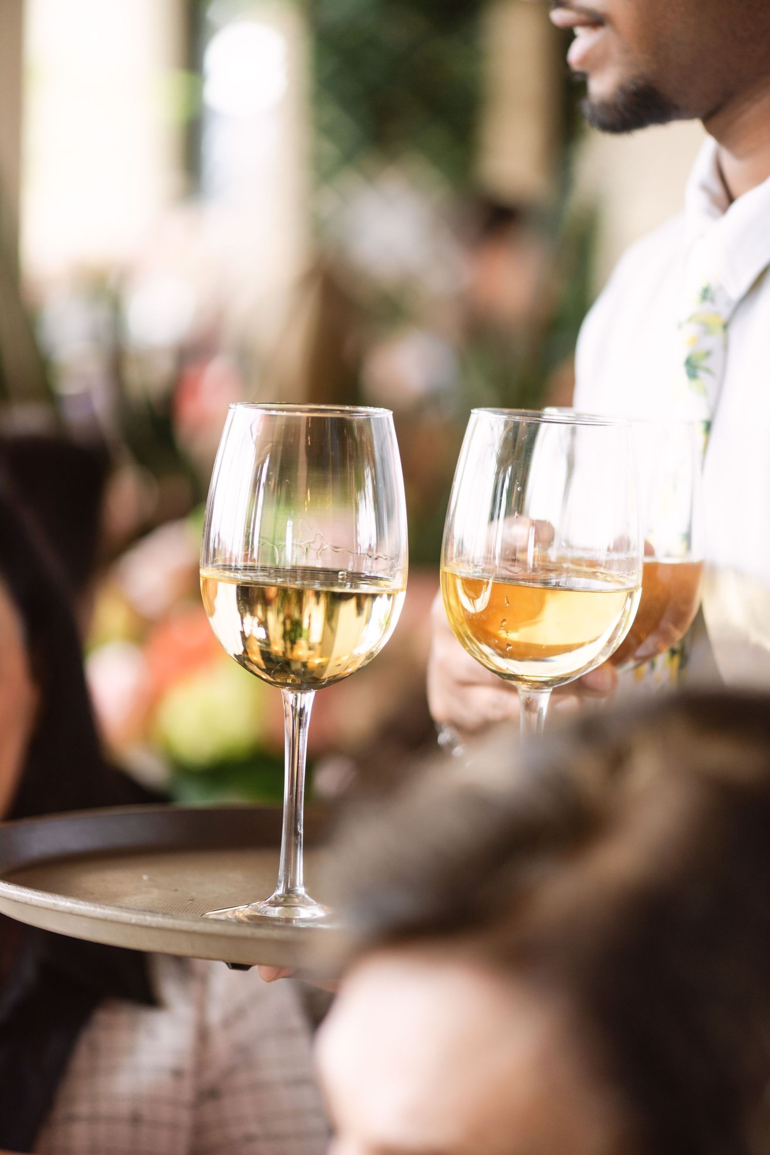 A waiter serving two glasses of white wine on a tray at an outdoor restaurant terrace.