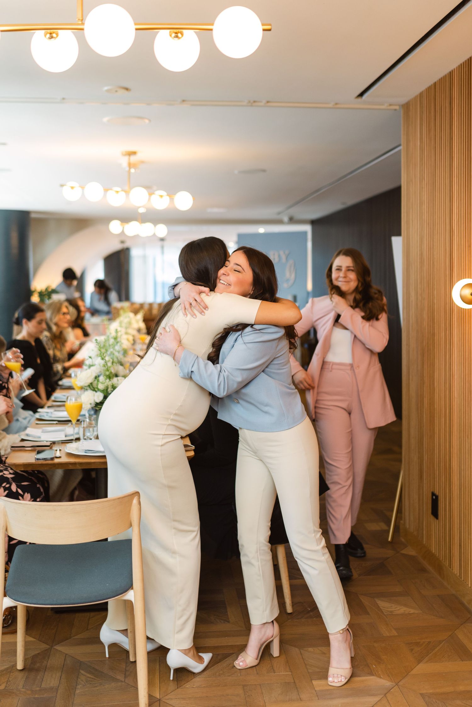 People in white and pastel outfits embrace and laugh together during a social gathering in a modern indoor space.