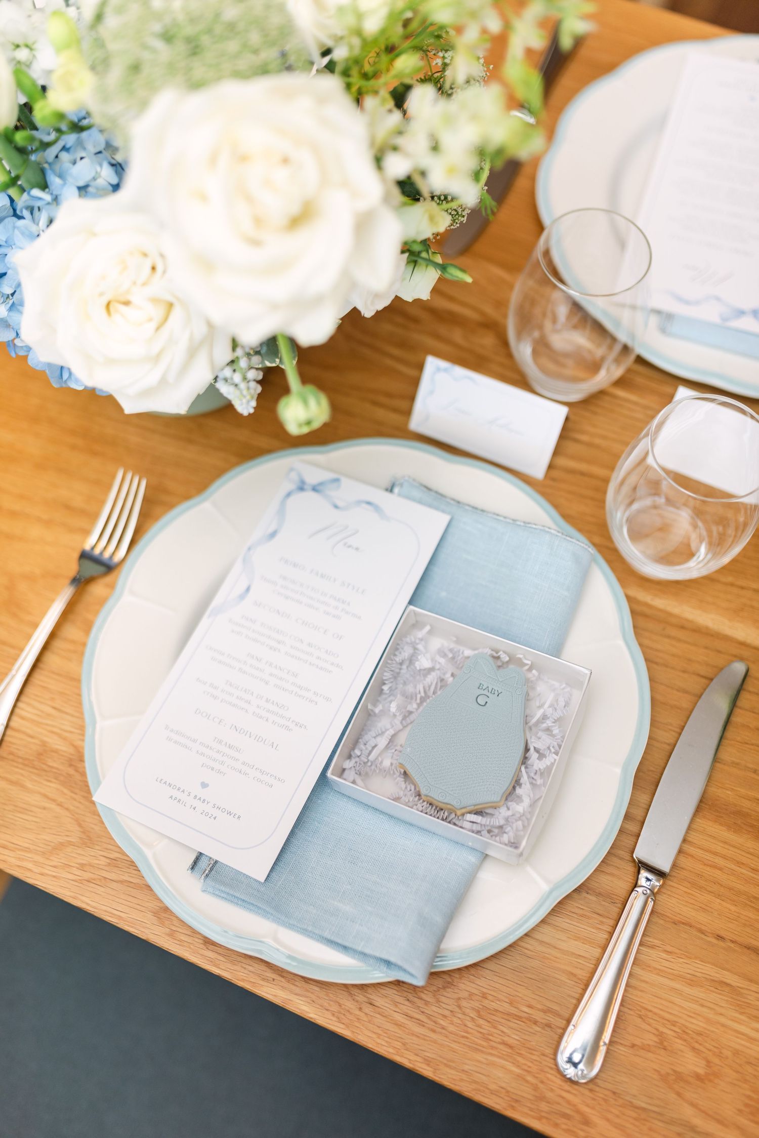 Elegant place setting with light blue napkin, white plate, and white floral centerpiece on wooden table.