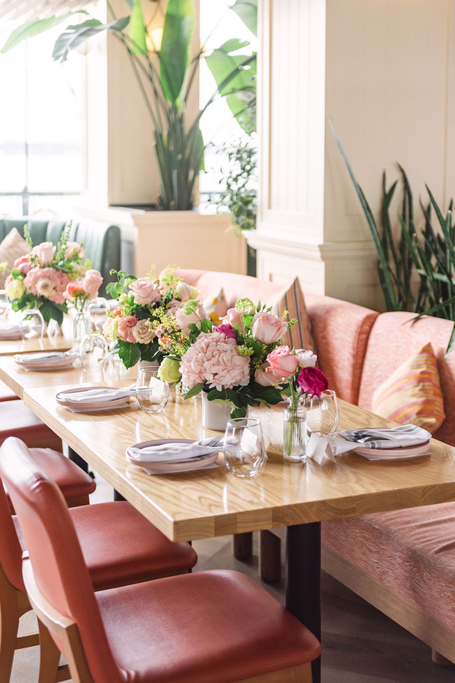 Elegant dining table with pink floral centerpieces, red leather chairs, and lush greenery in a bright restaurant setting.