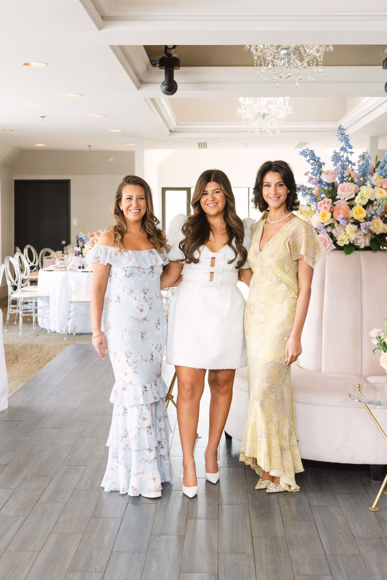 Three women posing together at a bridal shower, bride in white mini dress flanked by guests in floral gowns.