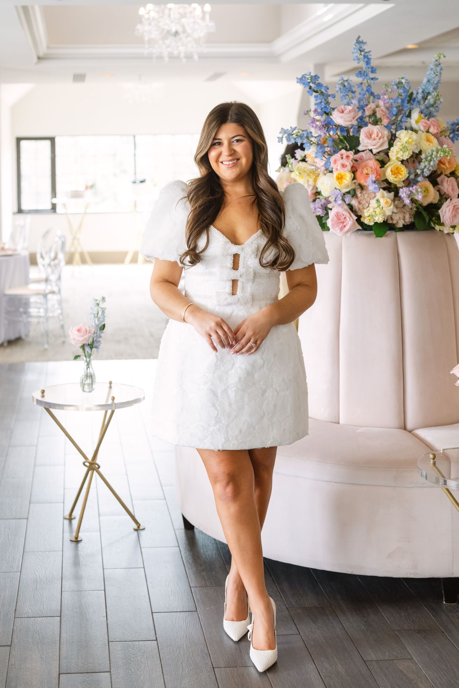 Smiling bride in white mini dress with puff sleeves and heels standing before a colorful pastel floral arrangement.