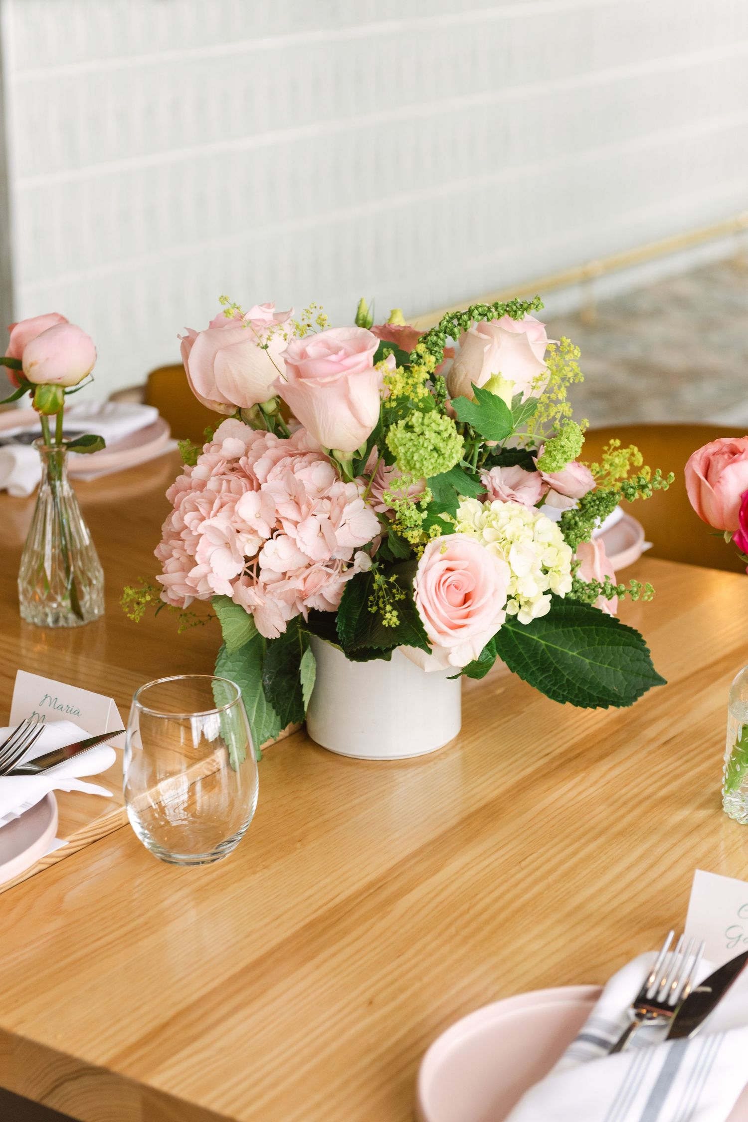Pink roses and hydrangeas in a white vase centerpiece on a wooden dining table with place settings.