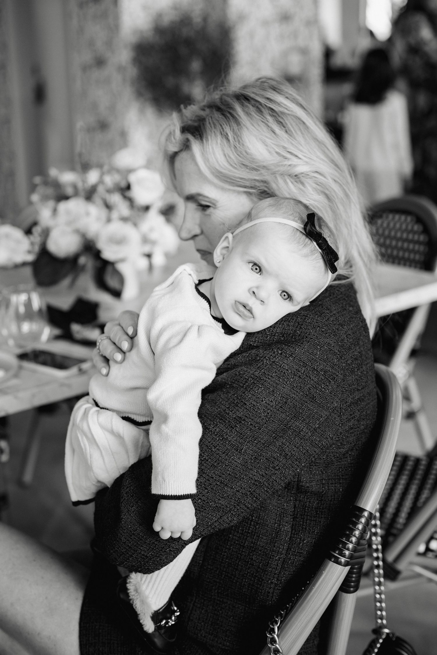 Black and white photo of a blonde woman tenderly holding a wide-eyed baby at an elegant floral event.