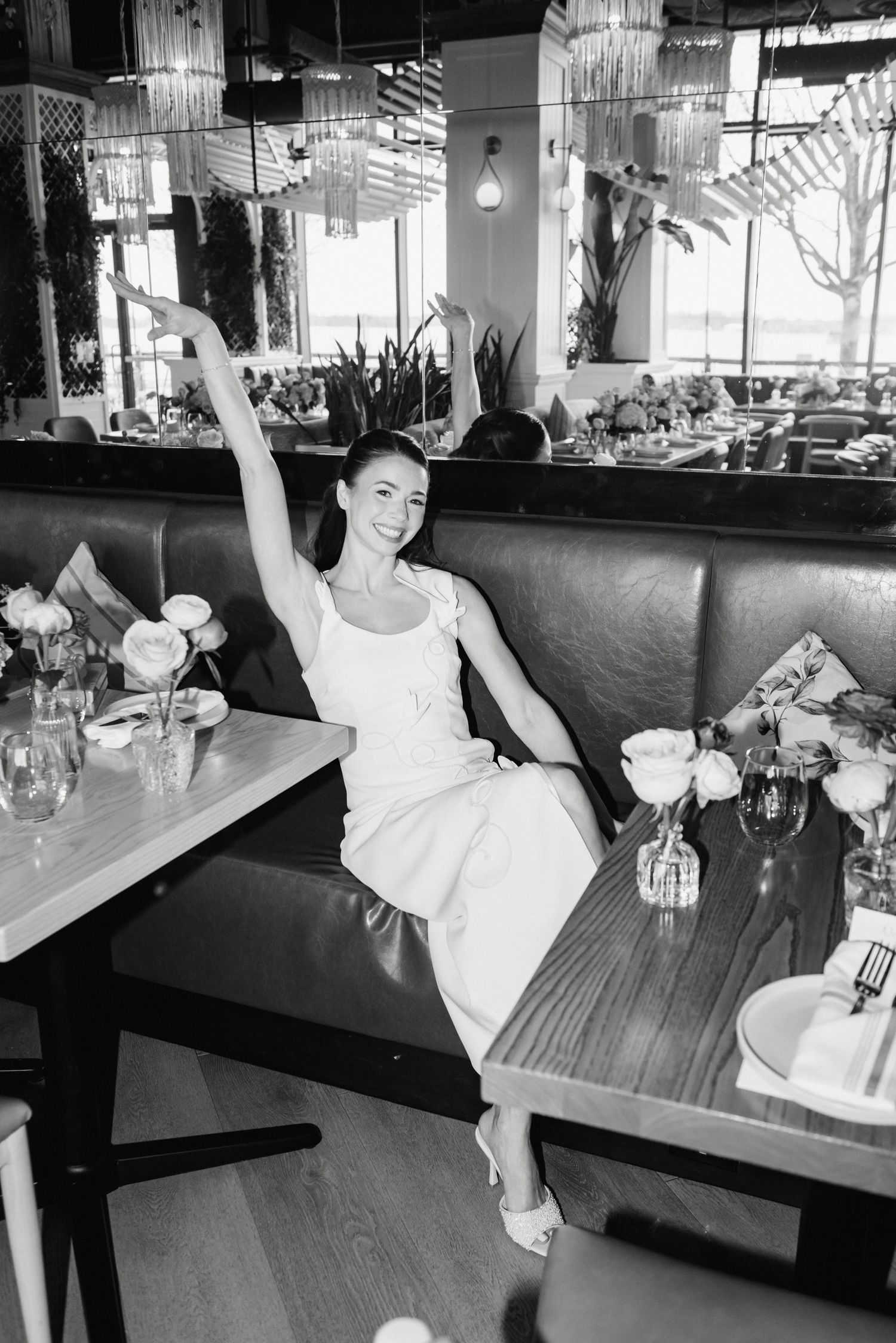 Black and white photo of a smiling woman in a white dress raising her arm in a stylish restaurant booth.