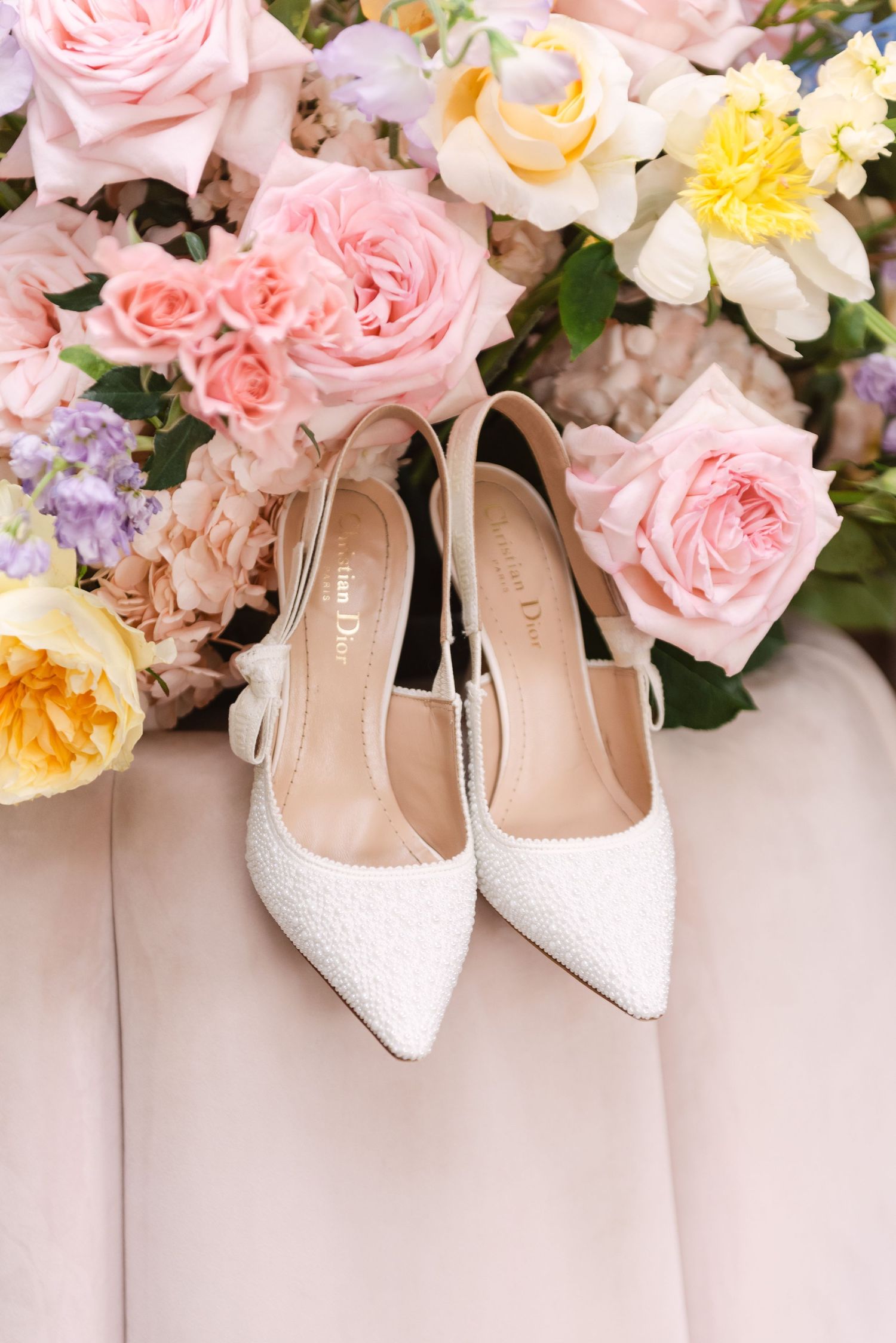 White pearl Christian Dior slingback heels displayed against a lush arrangement of pink, yellow, and lavender roses.