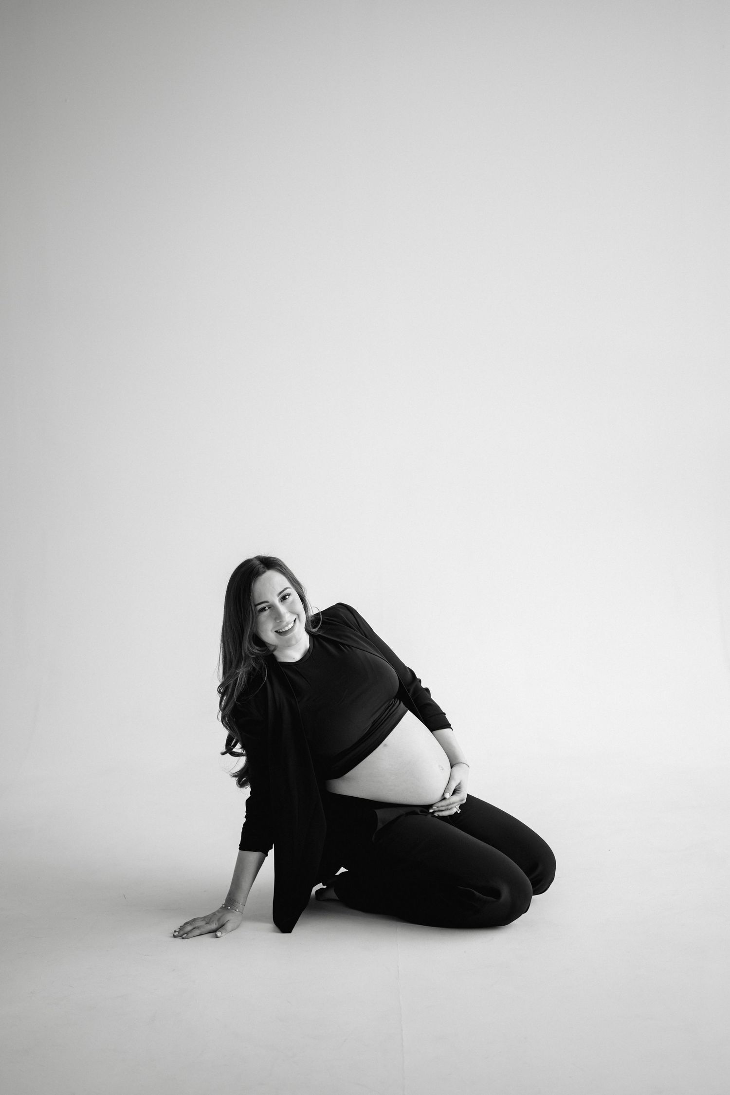 Artistic black and white maternity photo with a relaxed casual pose on a light studio backdrop.