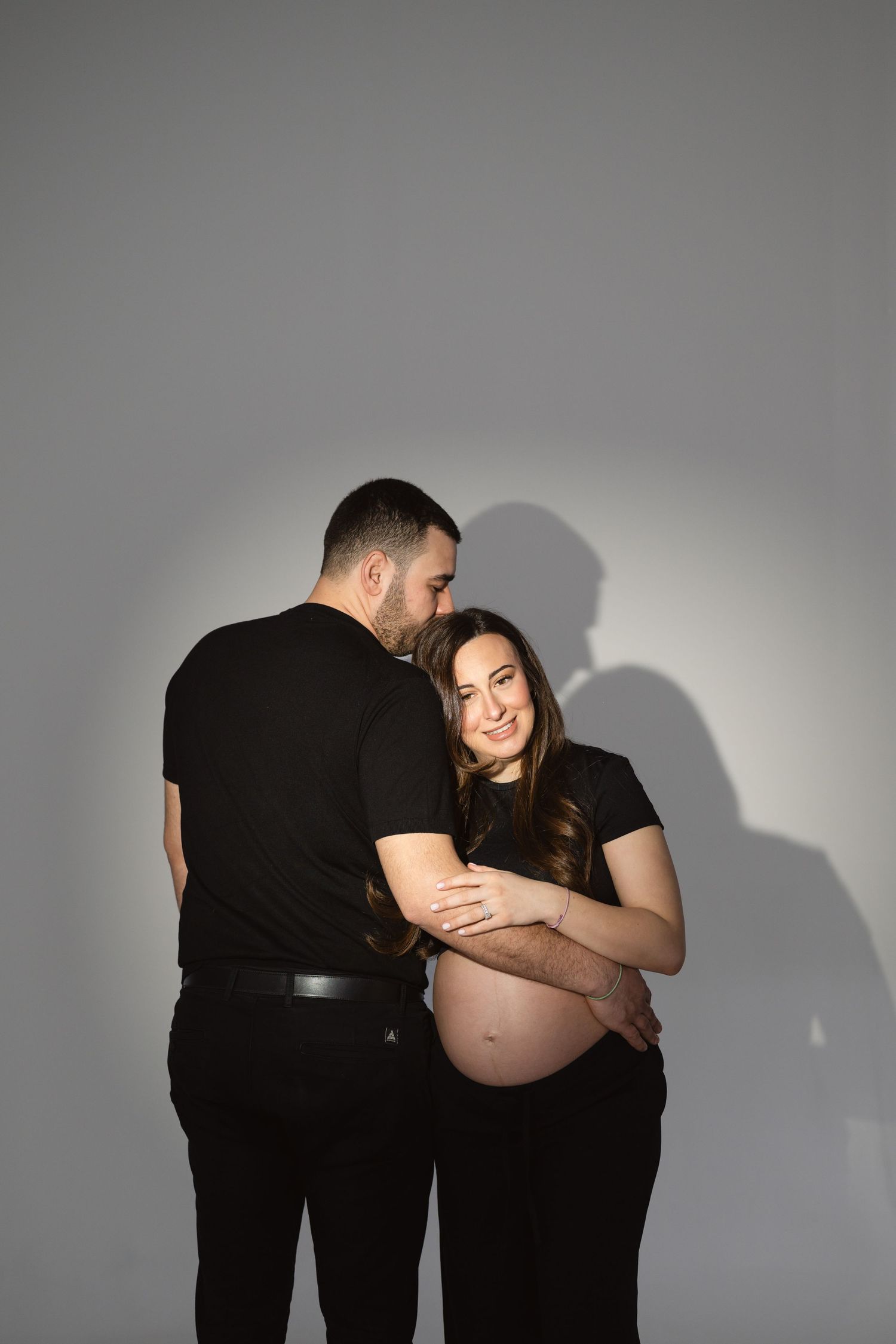 A series of intimate maternity photos showing a couple in black clothing against a light gray background with dramatic shadows.