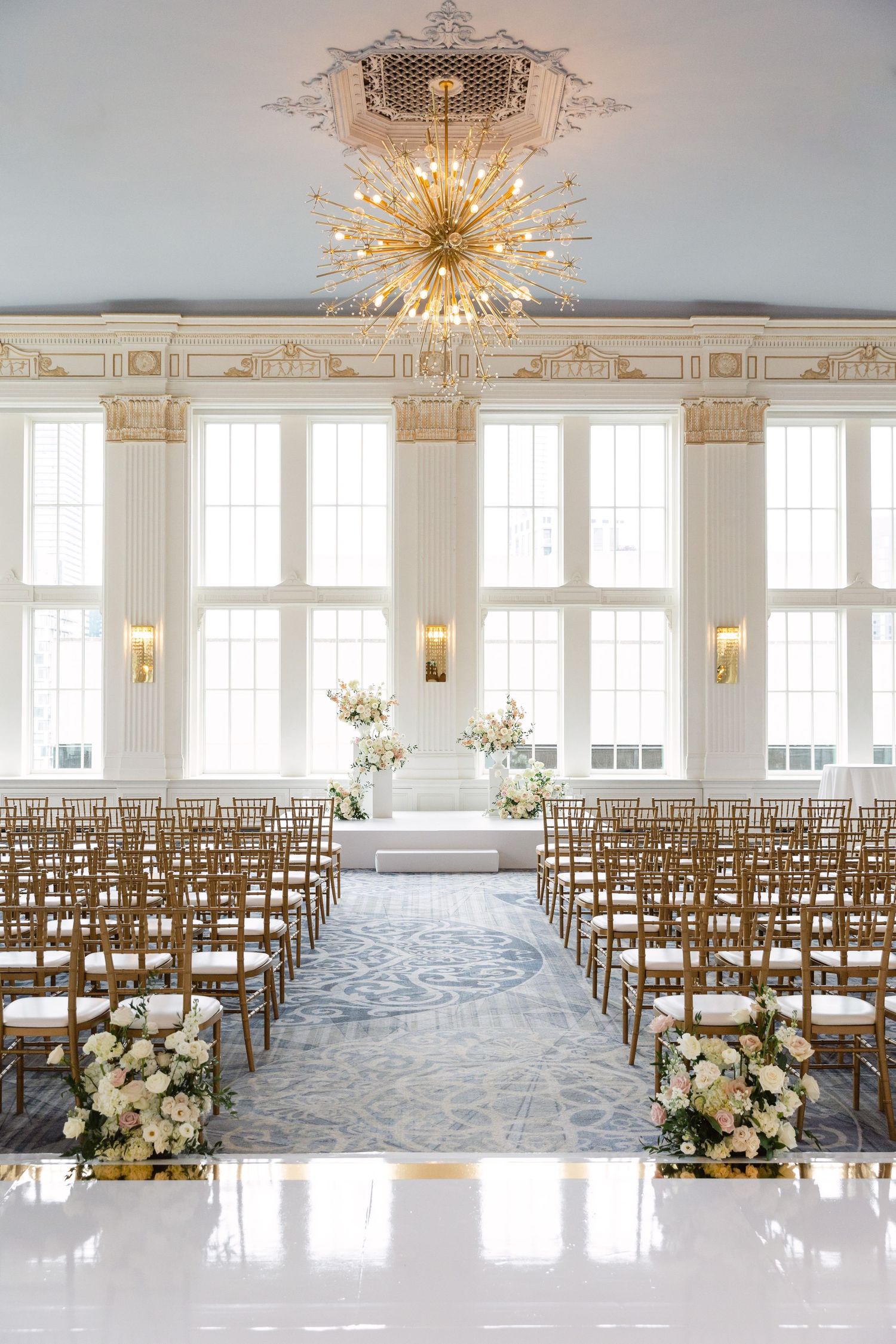 Elegant white wedding ceremony setup with wooden chairs and golden chandelier in a bright ballroom.