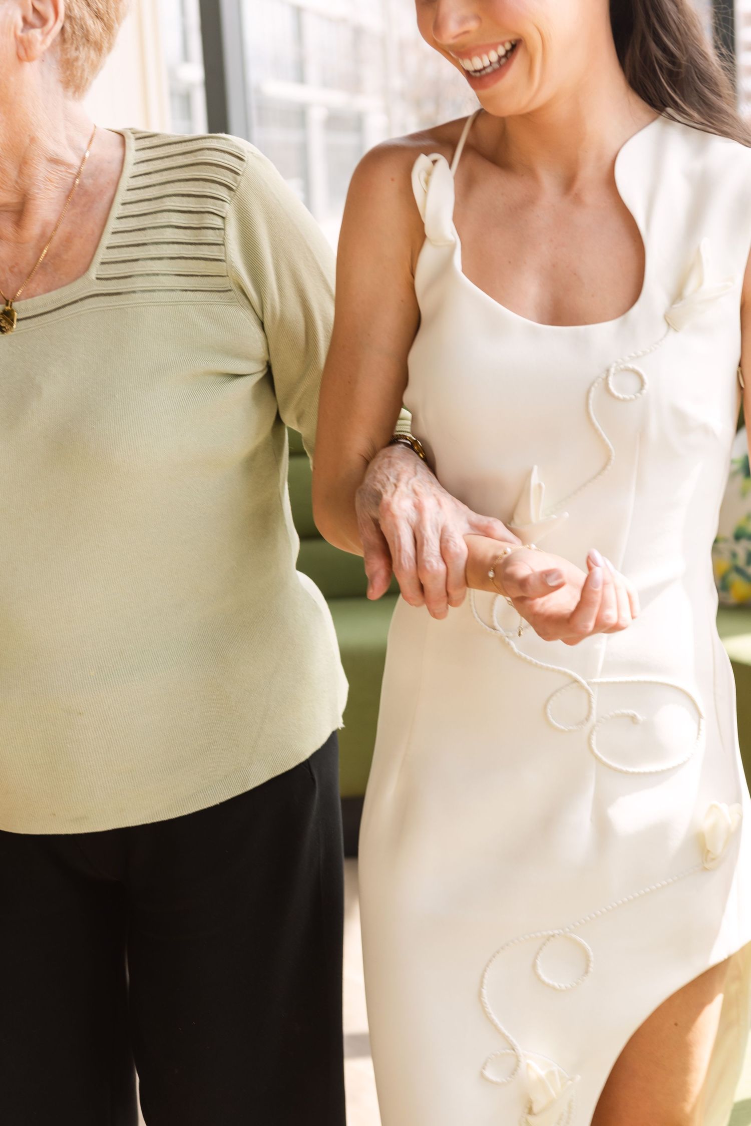 Elderly woman and young bride in white dress holding hands, sharing a tender moment together.