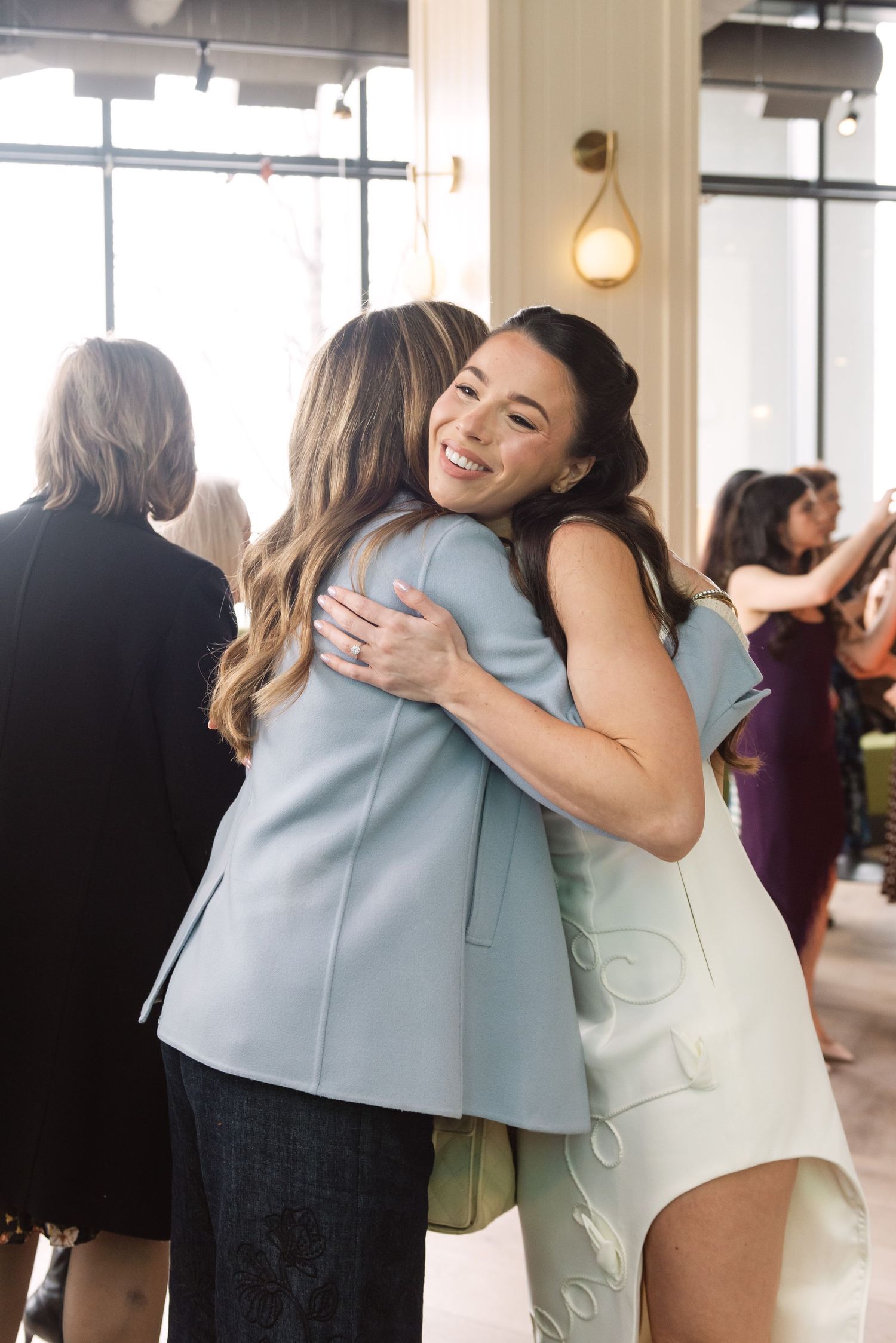 Two women in pastel blazers share a warm hug and smile at an elegant indoor social gathering.