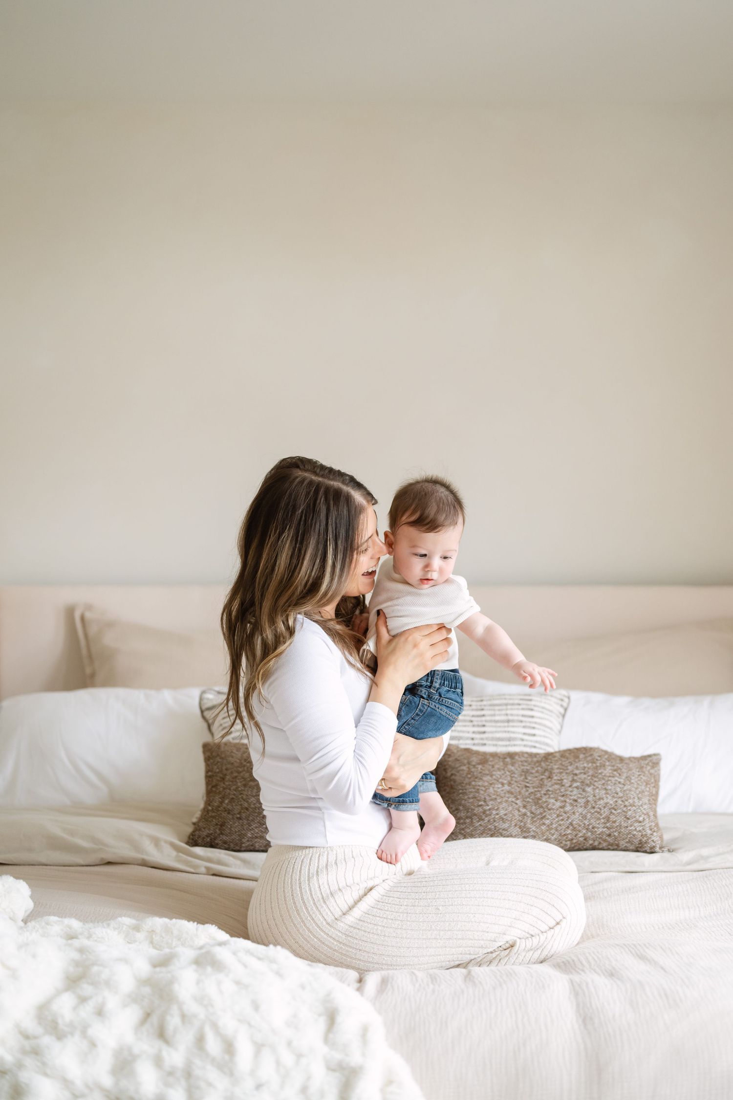Mother and baby share tender moment while sitting on white bedding in bright minimalist bedroom setting.