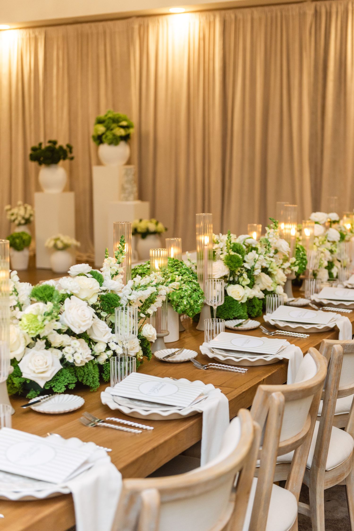Elegant wedding reception table with white floral centerpieces, wooden tables, and cream-colored chairs in a banquet hall.