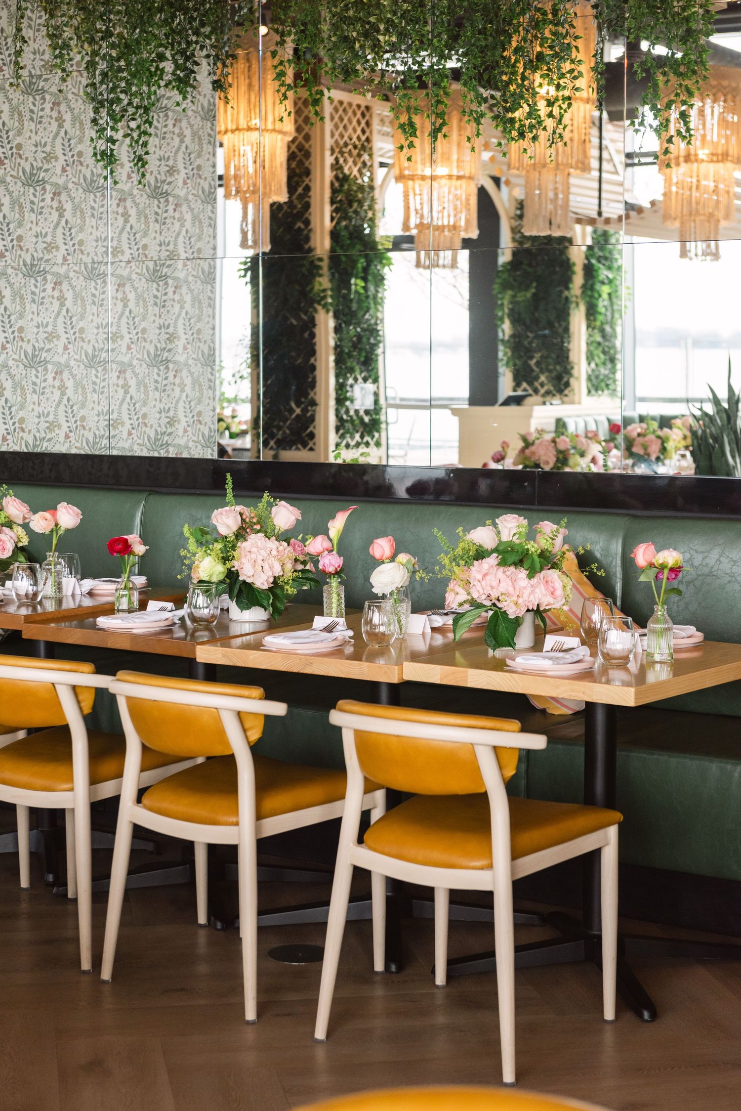 Elegant restaurant dining setup with yellow velvet chairs, pink floral centerpieces, green living walls, and gold pendant lights.