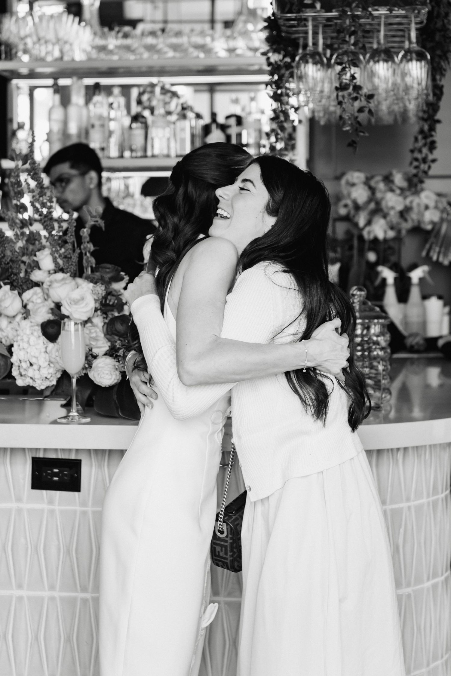 Two women in white dresses share a joyful embrace at a bridal shower bar in black and white photo.