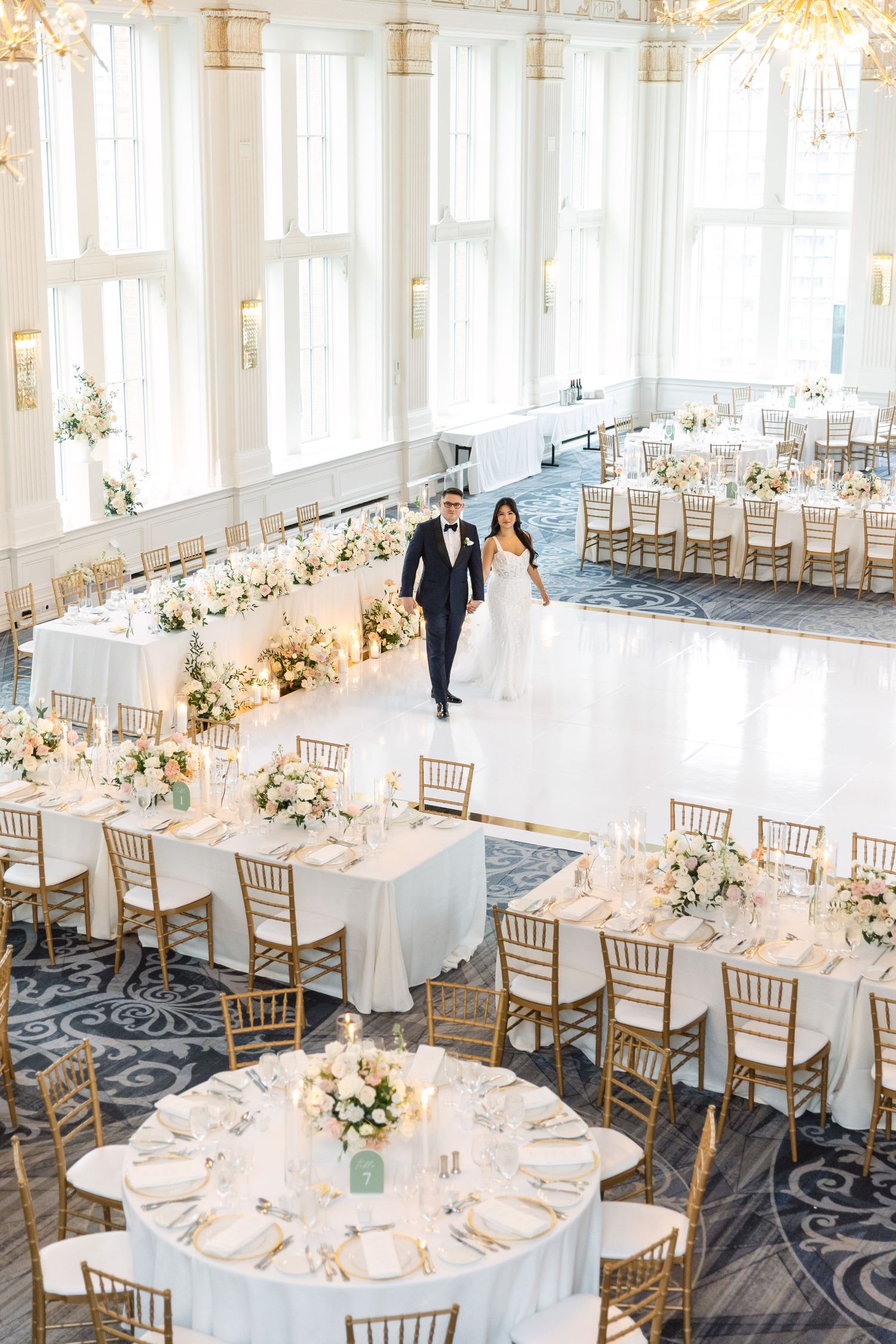 Elegant wedding reception setup with white tables and gold chairs under crystal chandeliers.